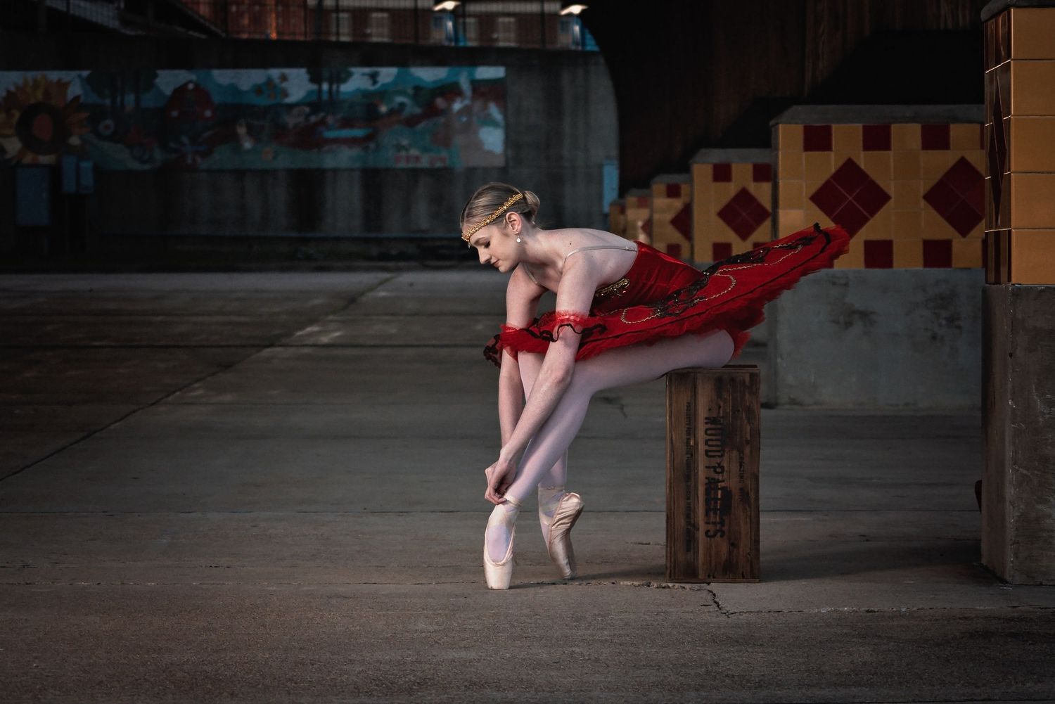 Ballerina in red tutu adjusts pointe shoes on wooden post in urban setting.