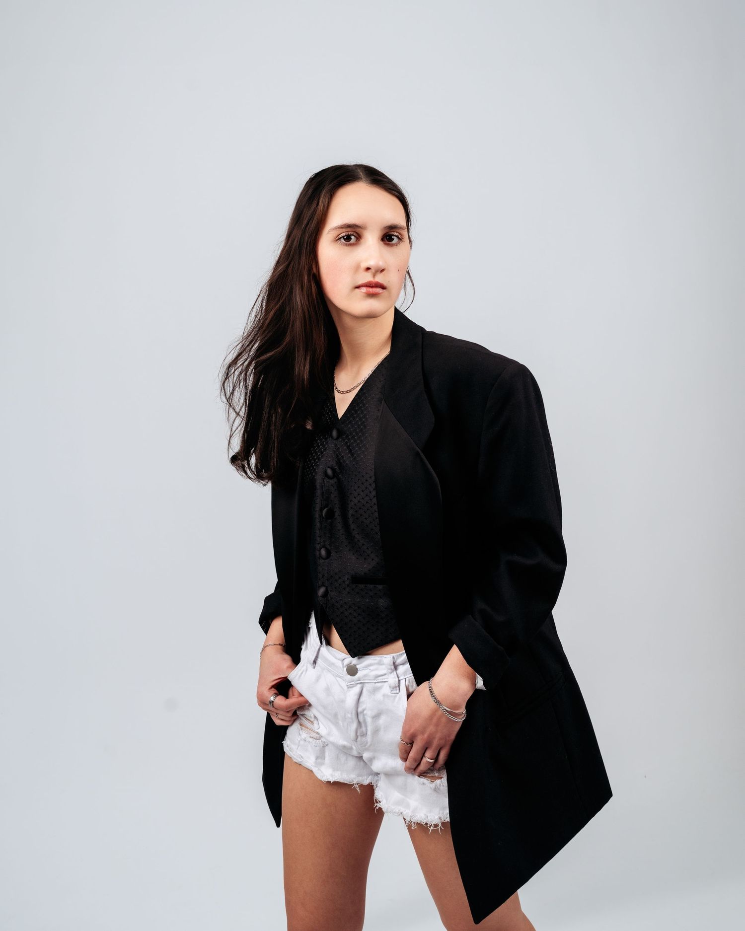 A young woman with long dark hair poses in a black blazer and white distressed shorts against a light background.