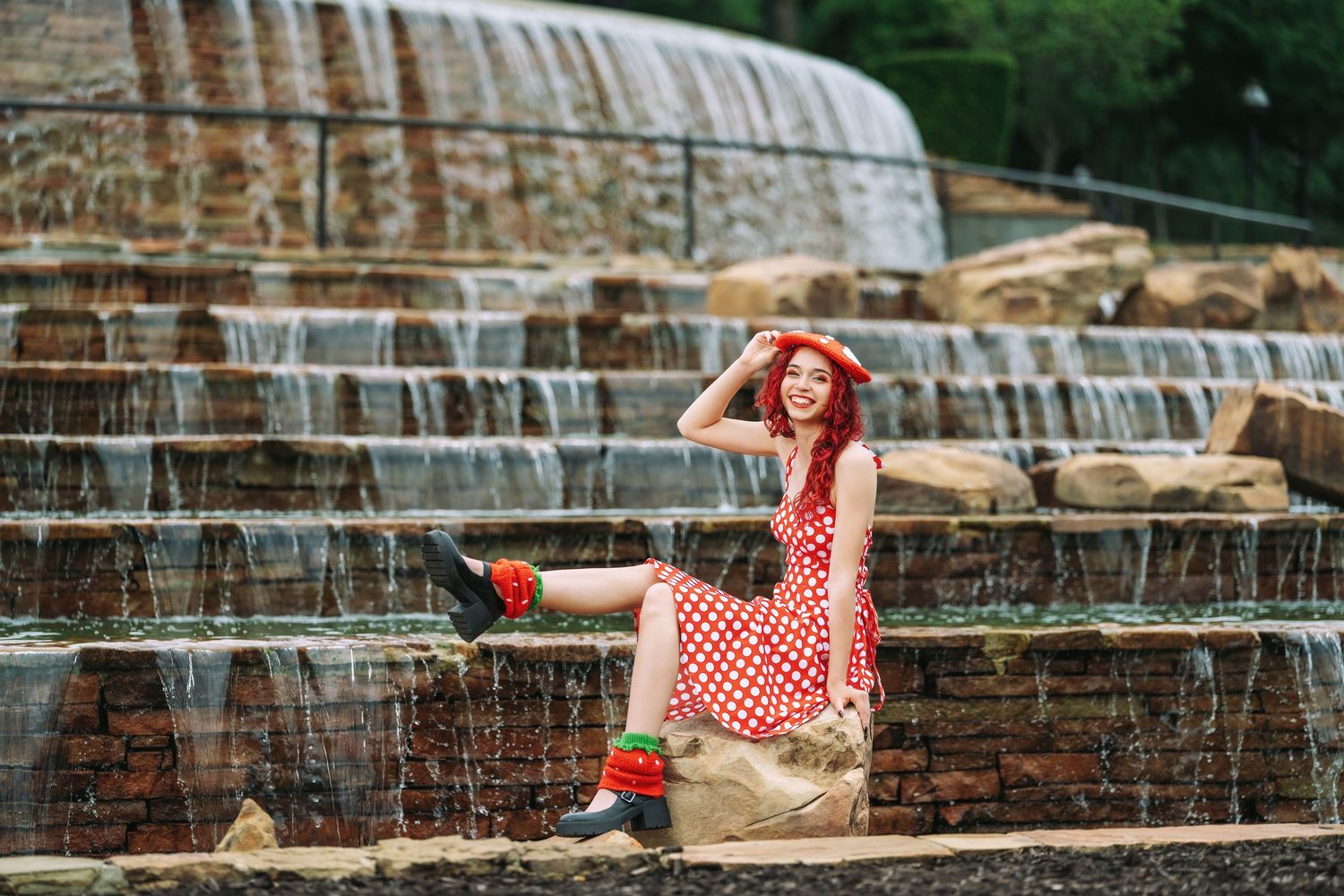 Red-haired woman in polka dot dress poses on stone steps beside cascading waterfall fountains outdoors.
