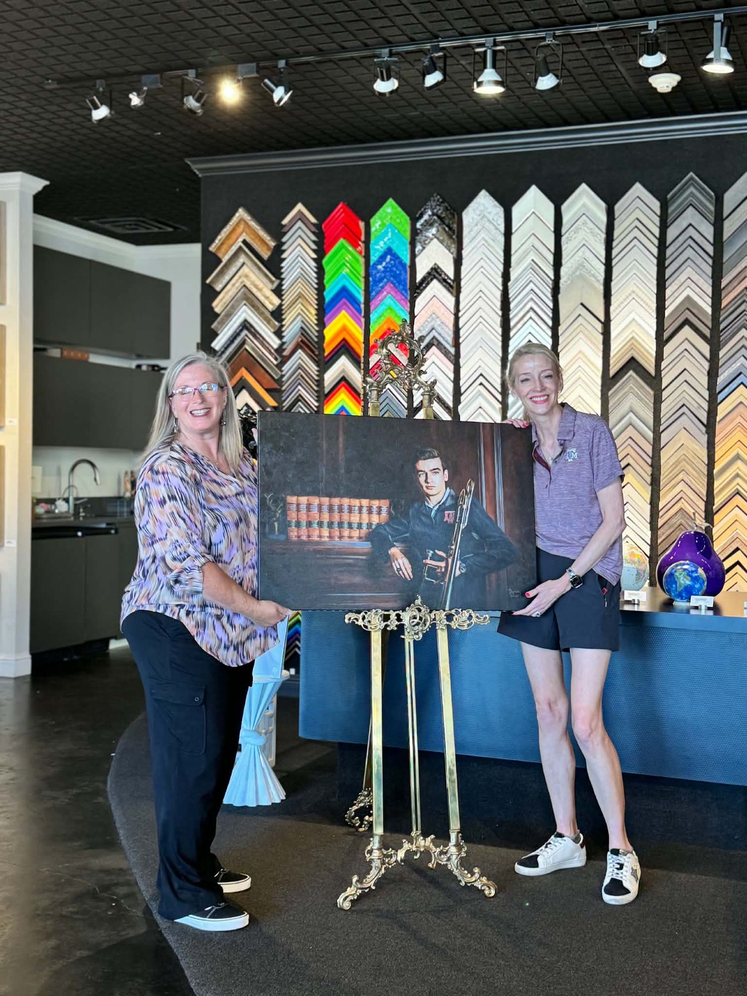 Two women display a framed portrait of a young man with guitar in a colorful frame shop.