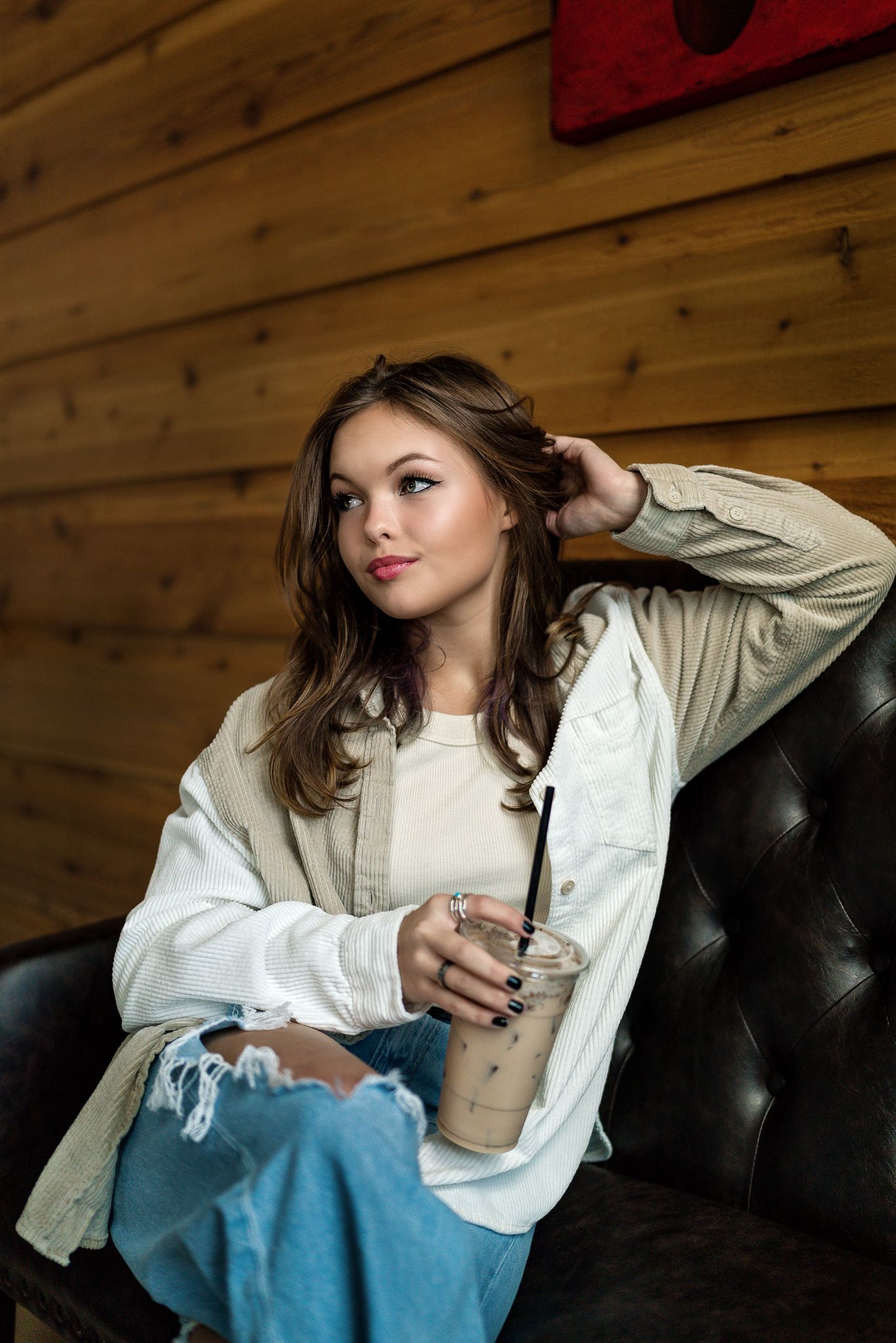 Young brunette woman relaxes in a leather chair holding an iced coffee drink against a wood wall.