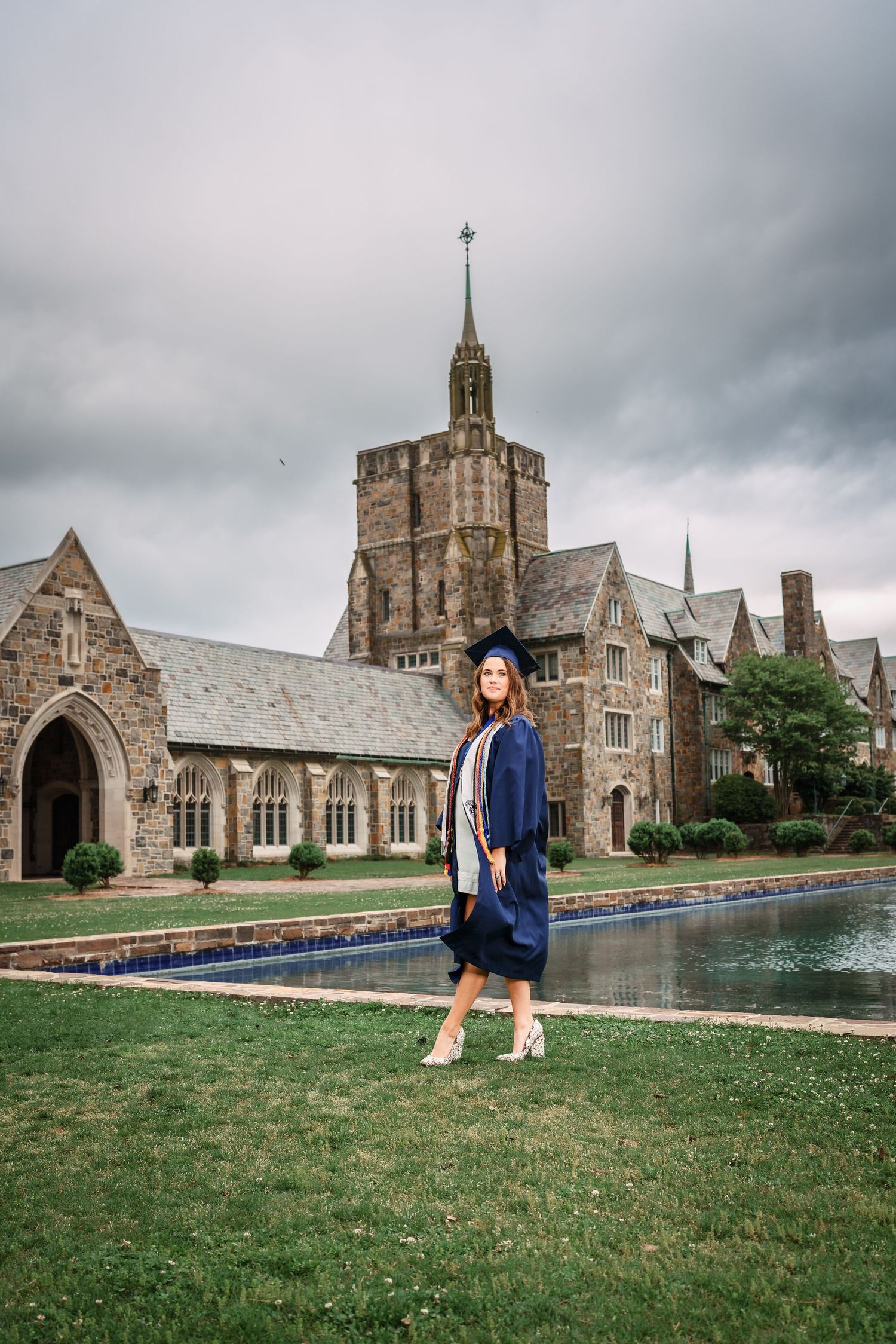 Graduate in blue cap and gown poses before historic stone university building with reflecting pool.