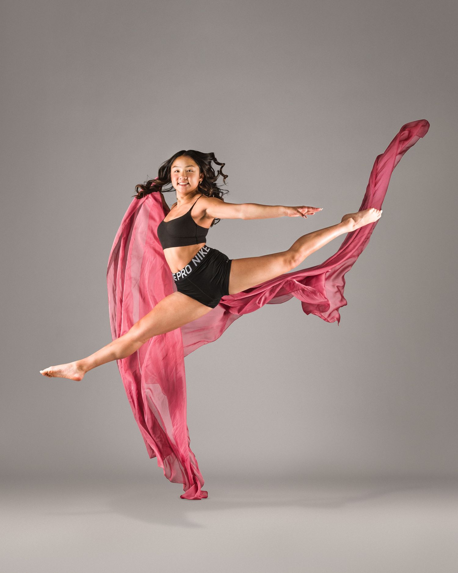 A dancer leaps gracefully in black athletic wear, holding flowing pink fabric against a gray studio background.