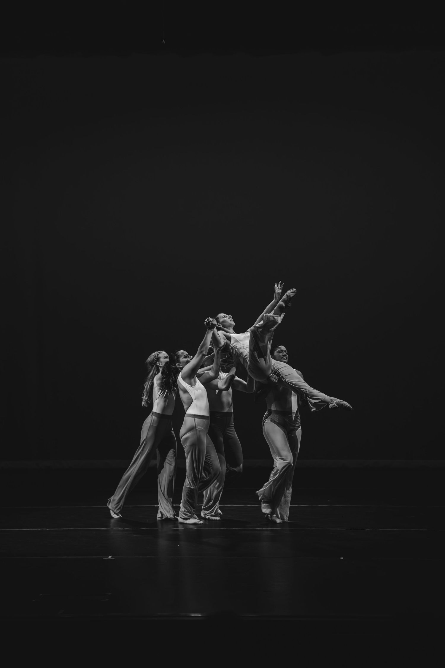 Black and white photo of contemporary dancers performing a group lift on a dark stage.