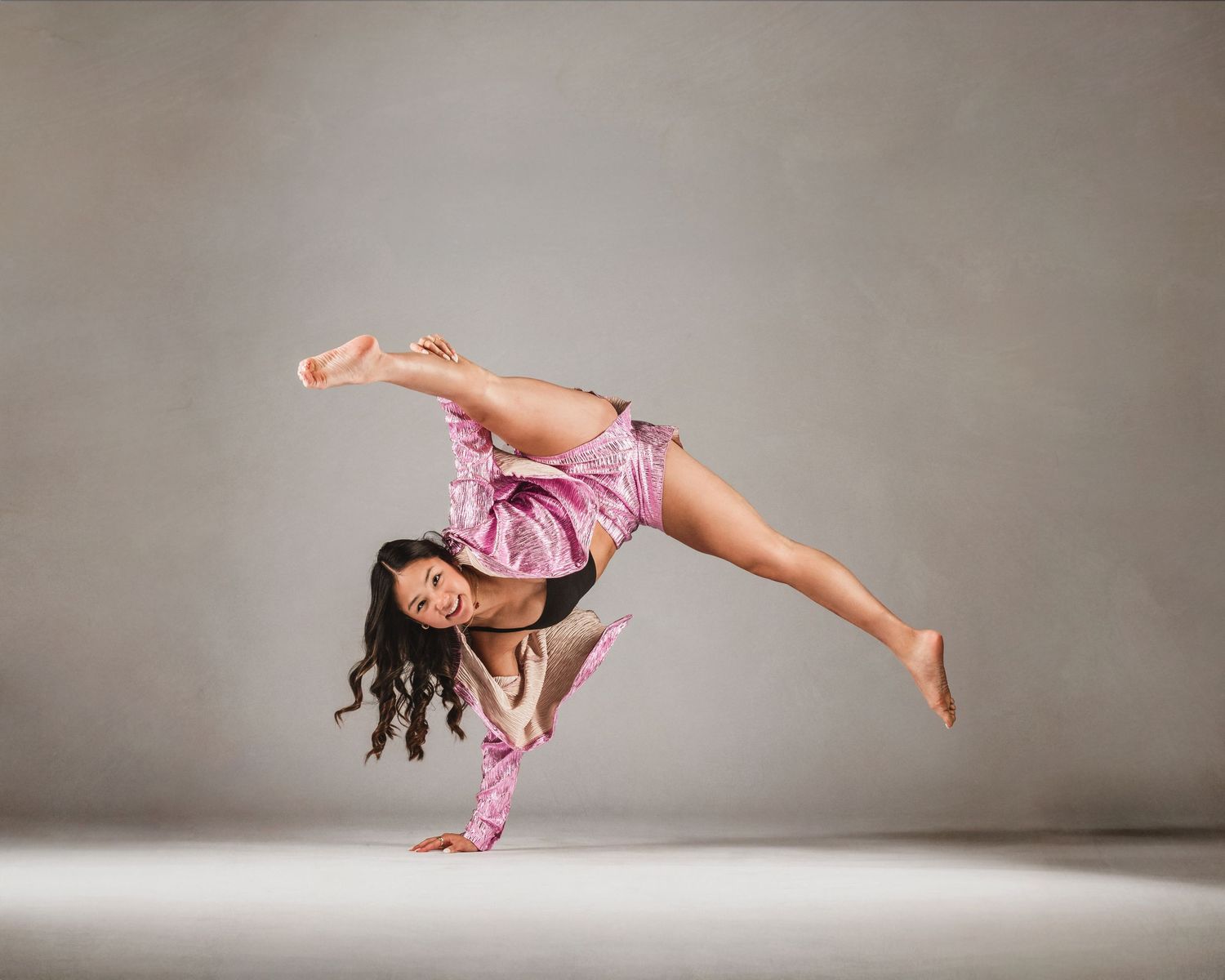 A flexible dancer in a pink outfit performs a one-handed handstand with legs split in a studio.