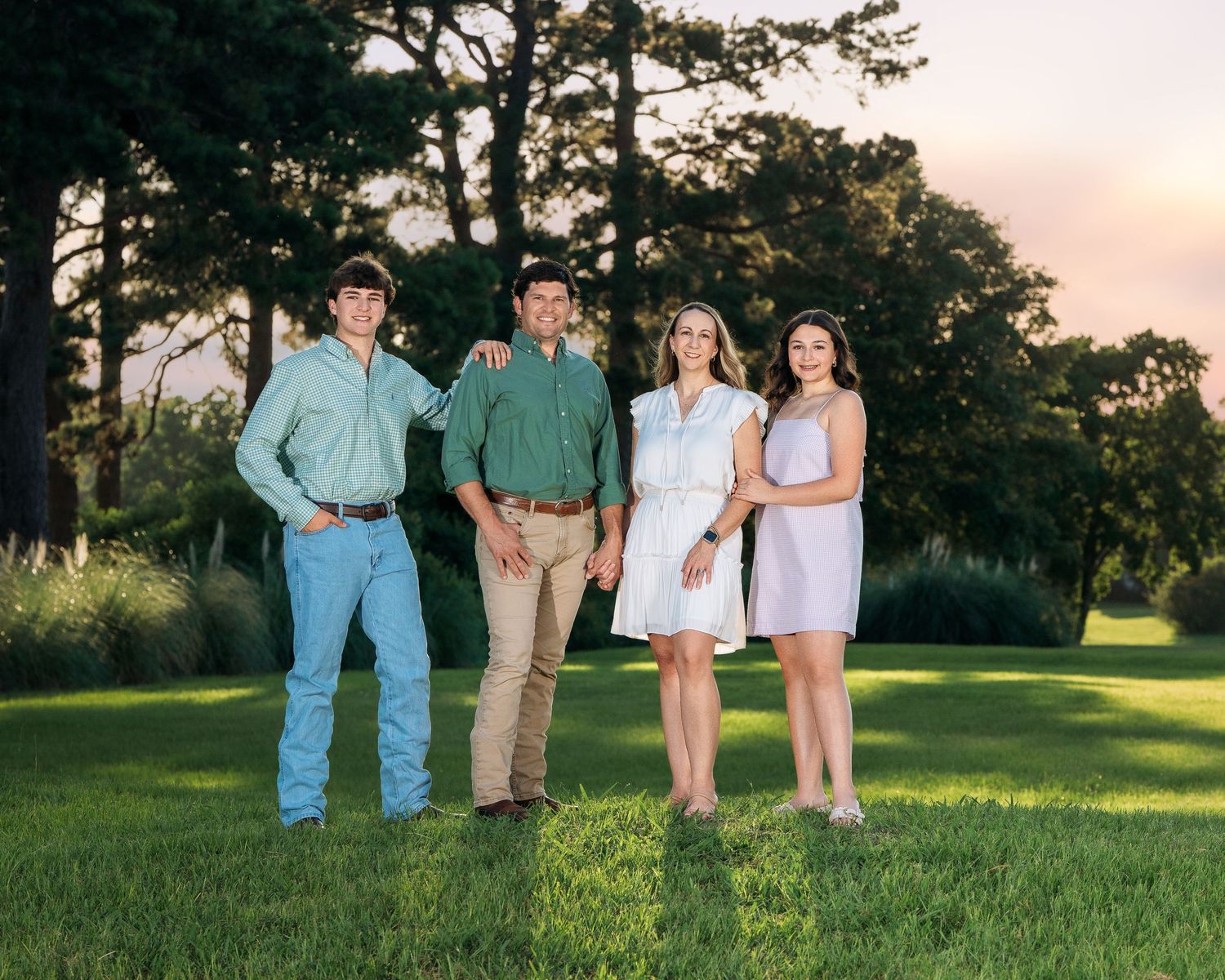 Family of four posing together on green lawn at sunset with tall trees in background.