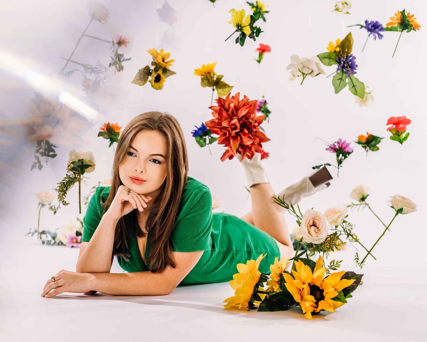 Young woman in green dress lying on floor surrounded by colorful artificial flowers in a bright studio setting.