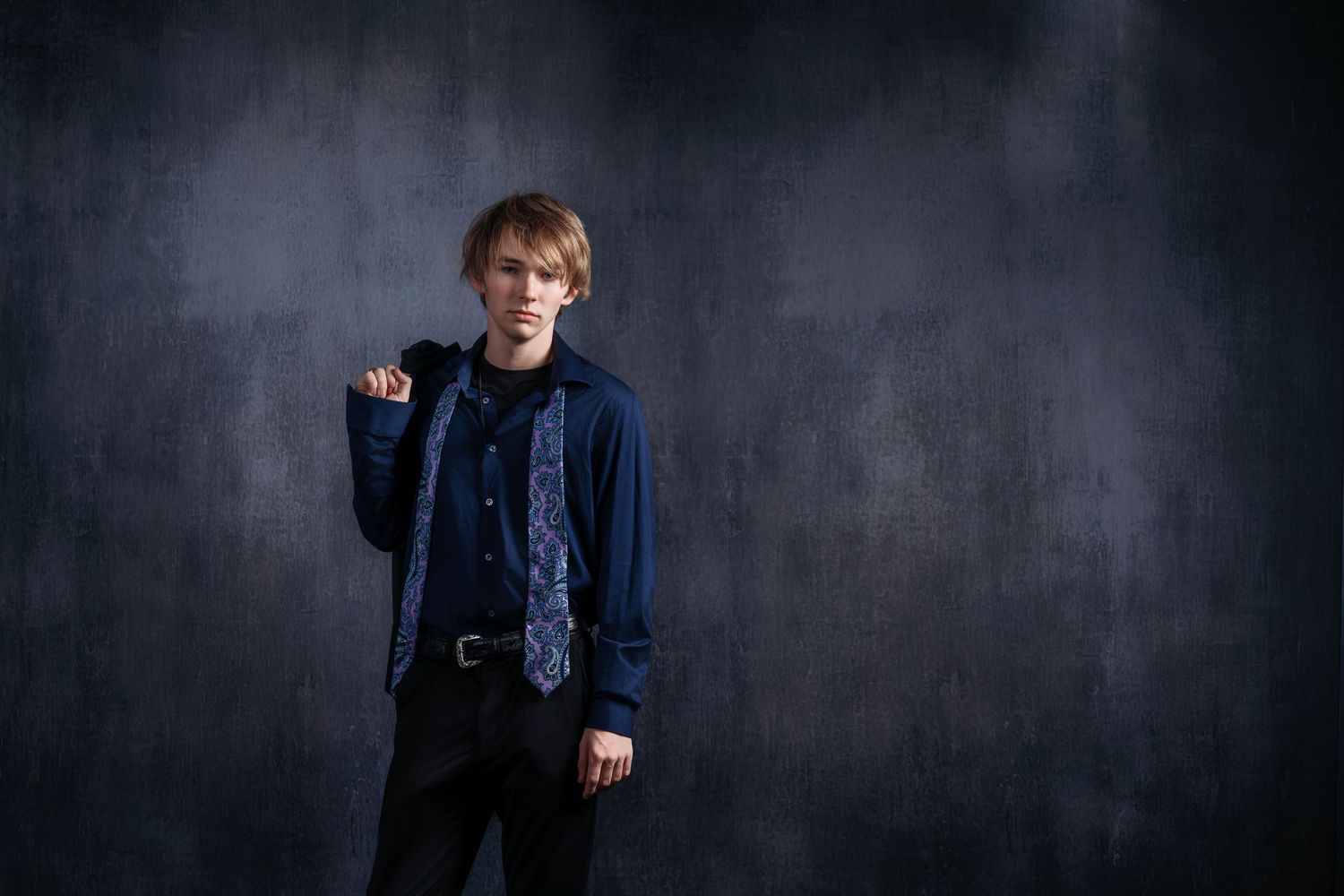 Young redhead male in blue shirt and tie poses confidently against dark gray studio background.