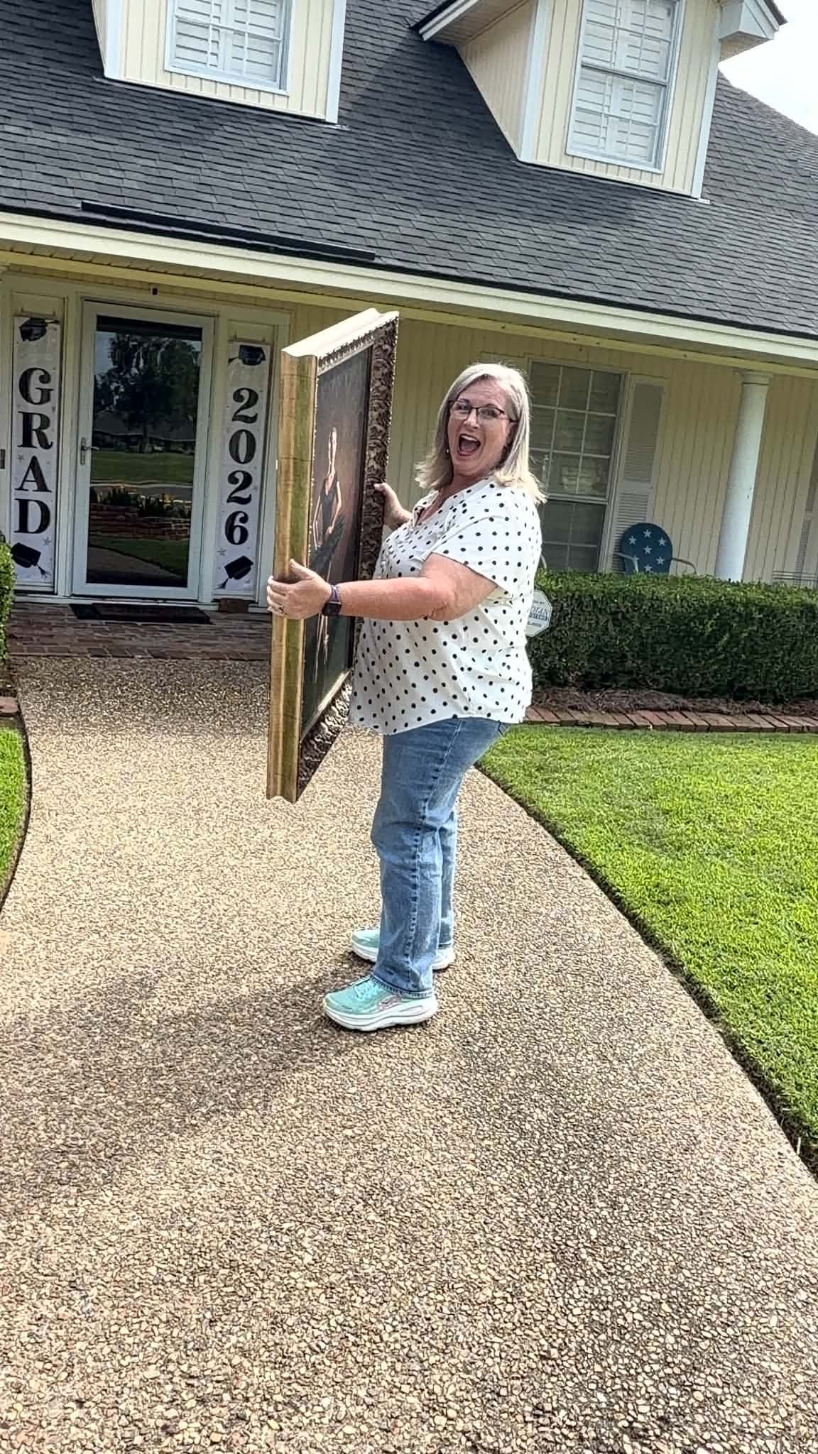 Happy woman holding a large framed portrait outside a home decorated with grad 2026 signs.