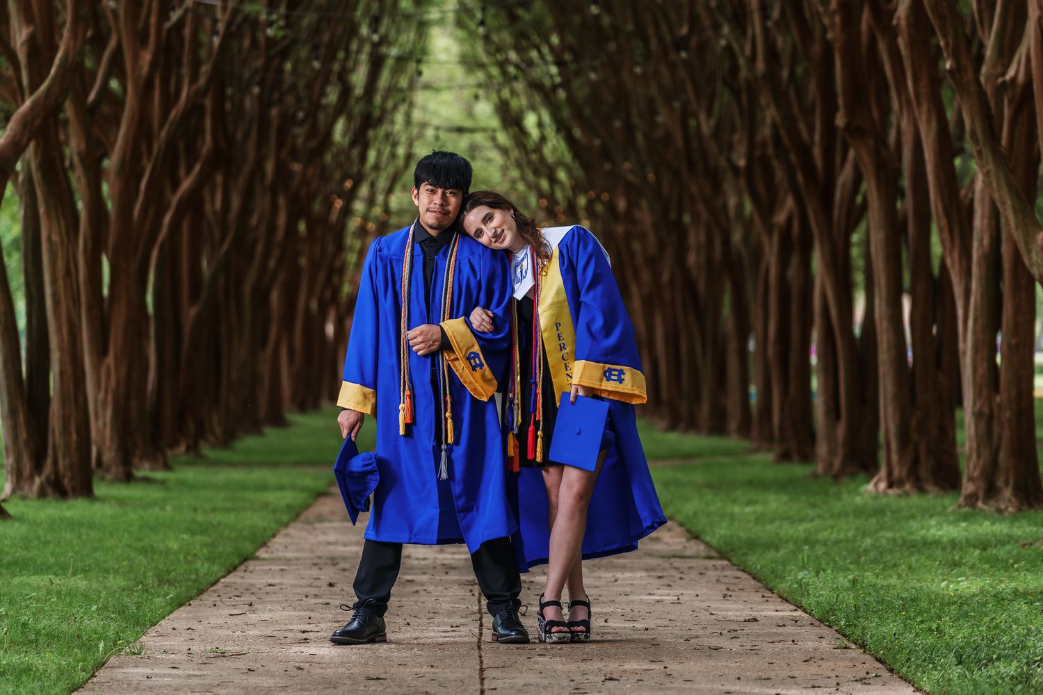 Two graduates in blue and gold regalia pose playfully on a tree-lined path.