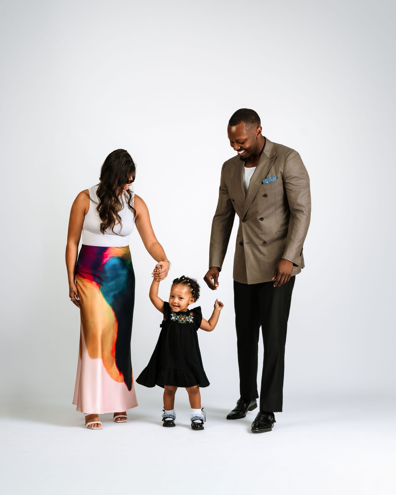 Family portrait of parents in elegant attire holding hands with their smiling toddler in black dress.