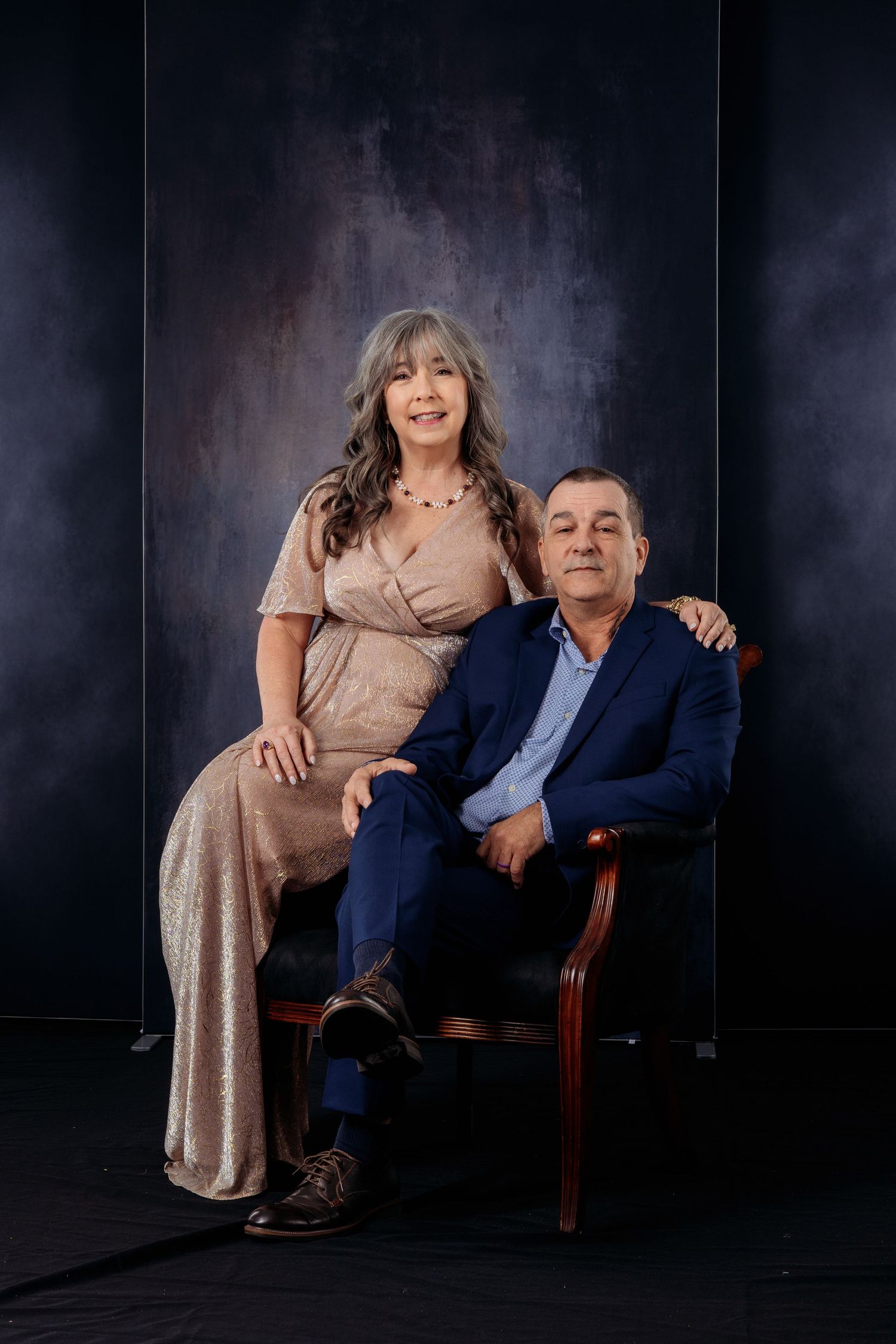 Elegant older couple poses together in formal attire against dark textured studio backdrop.