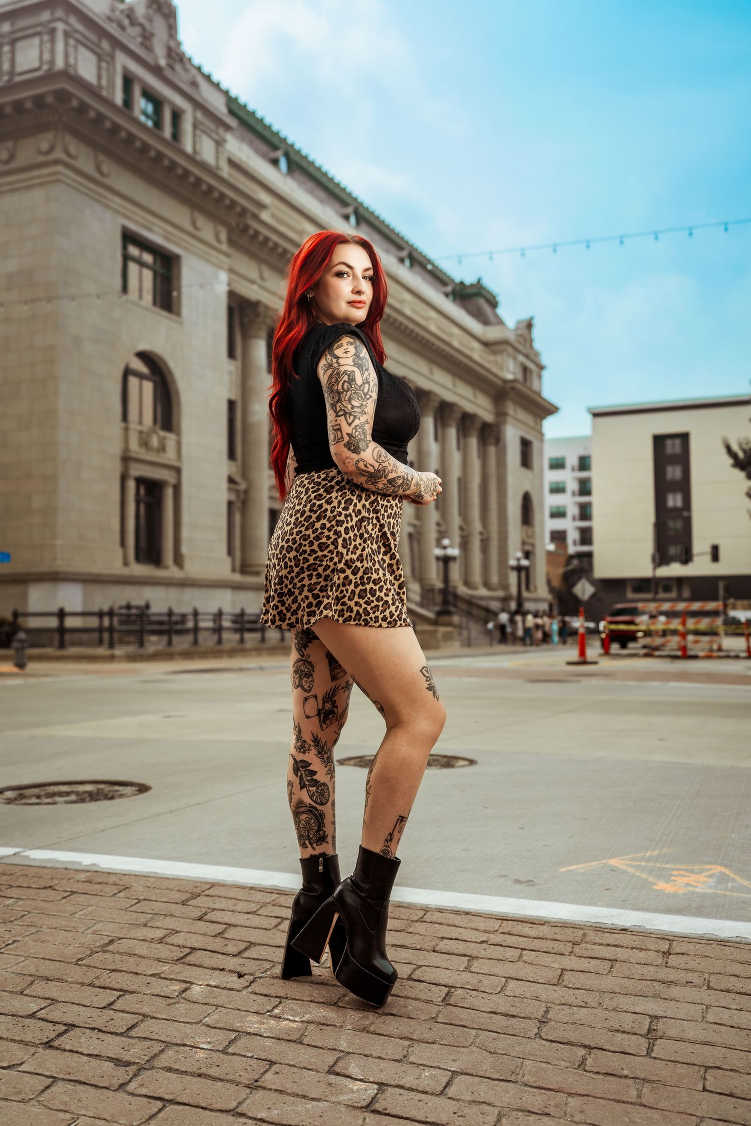 Tattooed woman with red hair poses in leopard skirt and black top on city street.