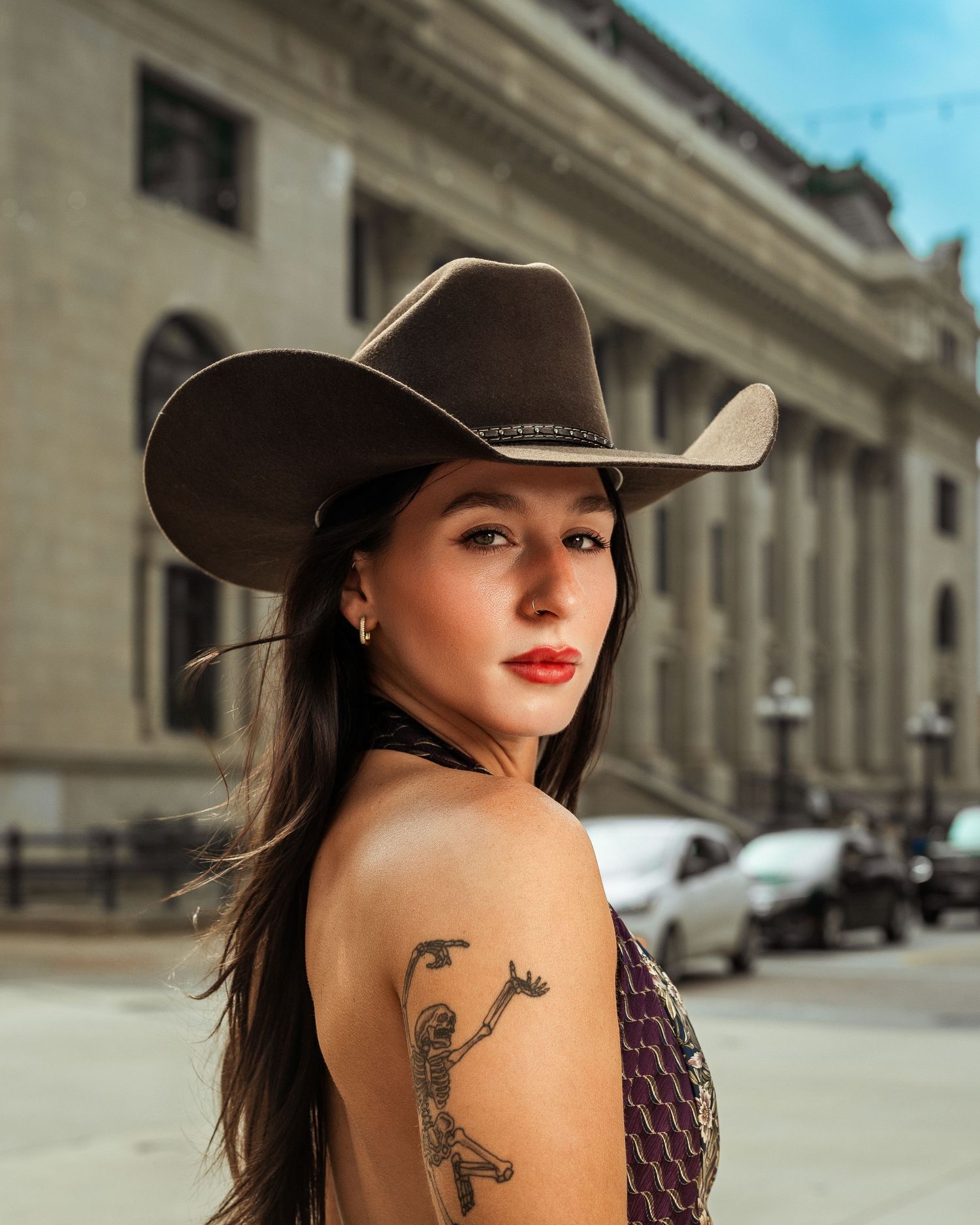 A young woman wearing a cowboy hat and shoulder tattoo poses on an urban street near a historic building.