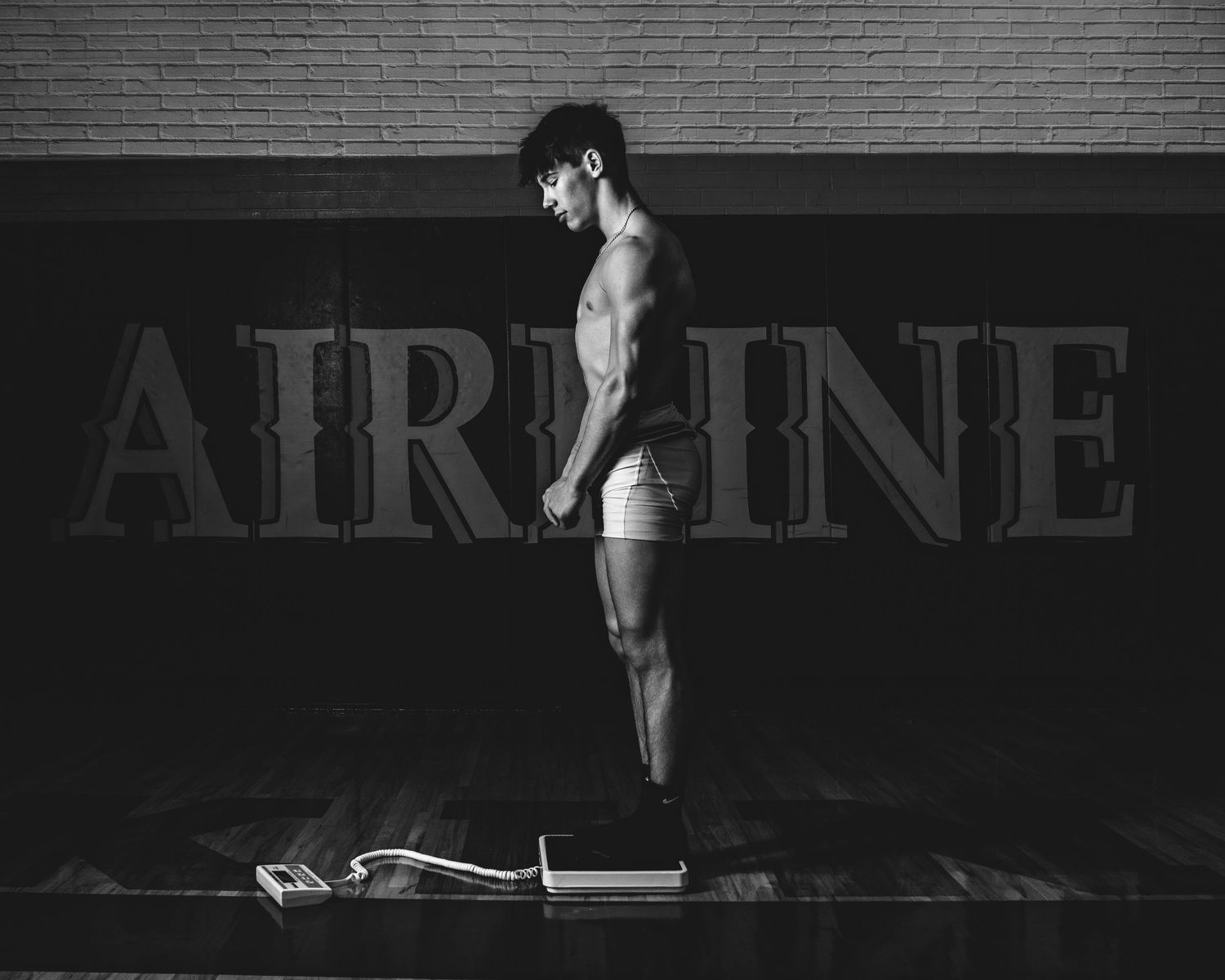 Black and white photo of a muscular man standing on a scale in front of an AIRLINE sign backdrop.