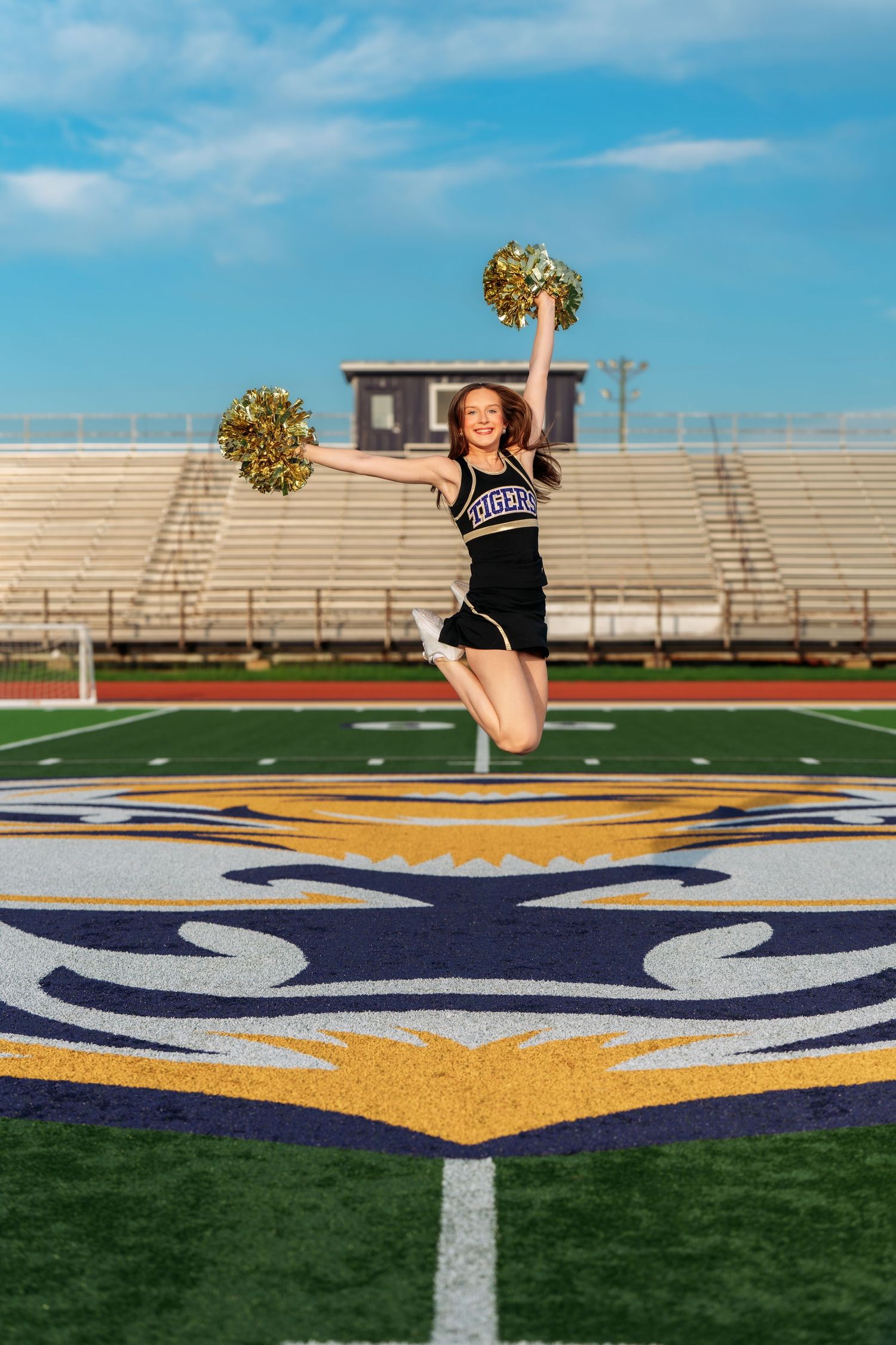 Cheerleader jumping with gold pom-poms on football field logo, wearing black uniform, stadium bleachers in background.