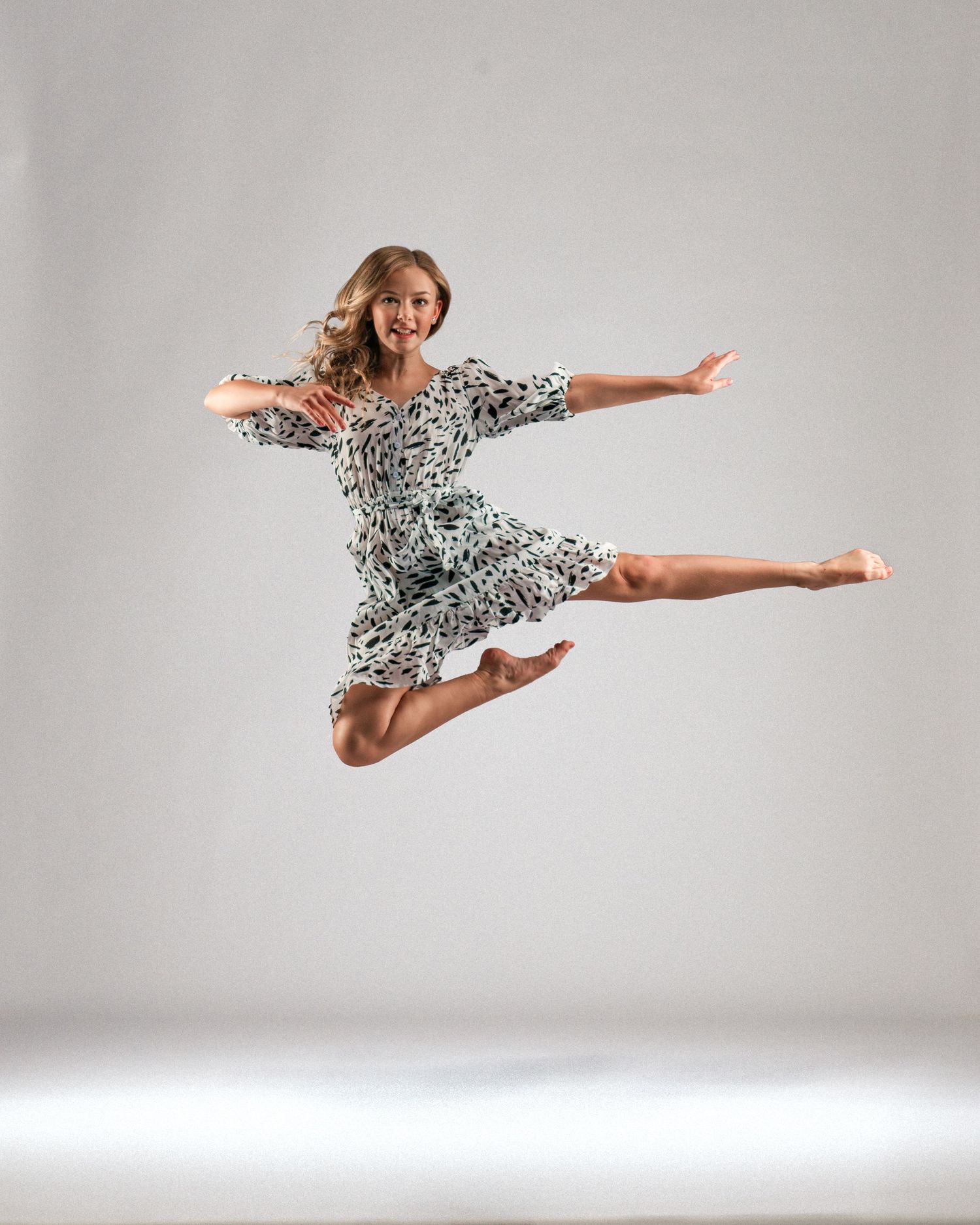 A dancer leaps gracefully in a floral dress, arms outstretched, against a light gray studio background.