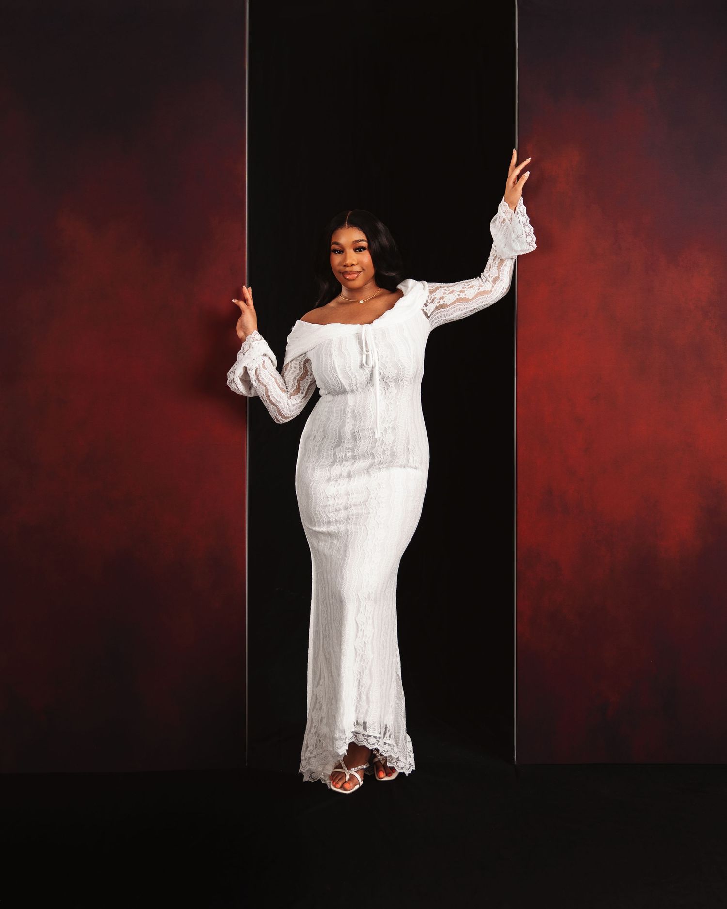 Woman in white lace off-shoulder gown poses elegantly against dramatic red and black backdrop.
