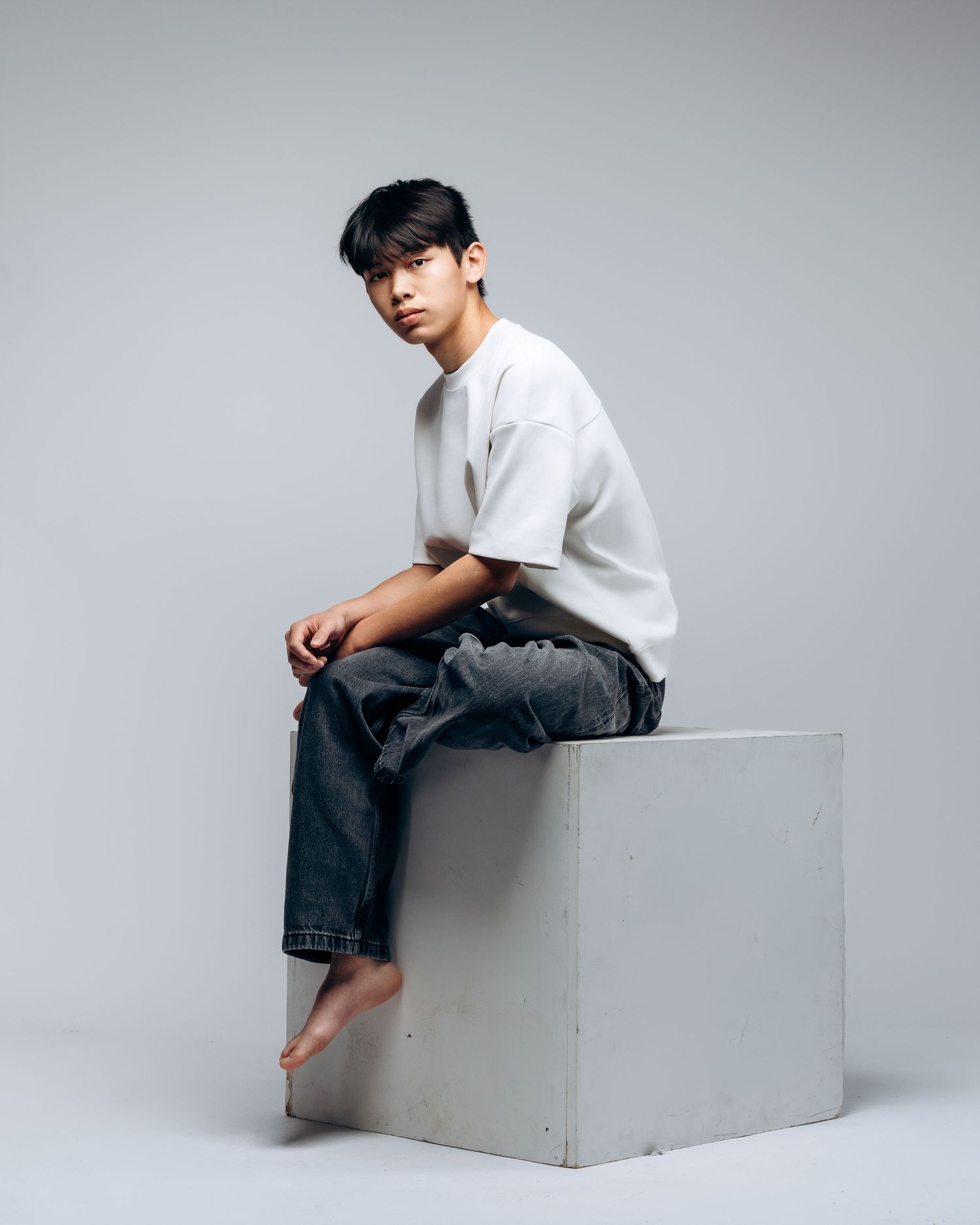 Young man in white t-shirt and dark jeans sits barefoot on a white cube block against a gray studio background.