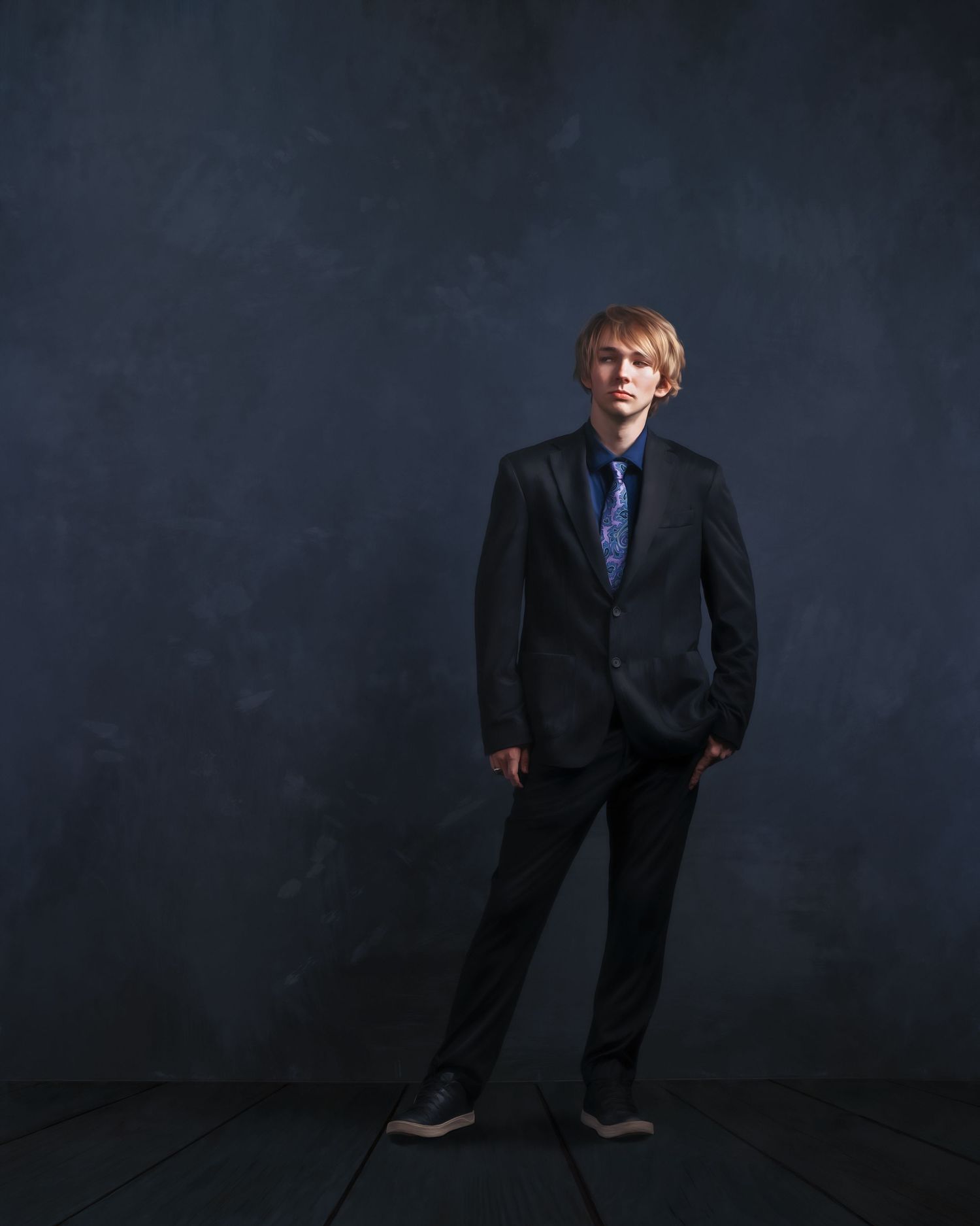 Young man in dark suit and blue tie poses confidently against a moody blue-gray studio backdrop.