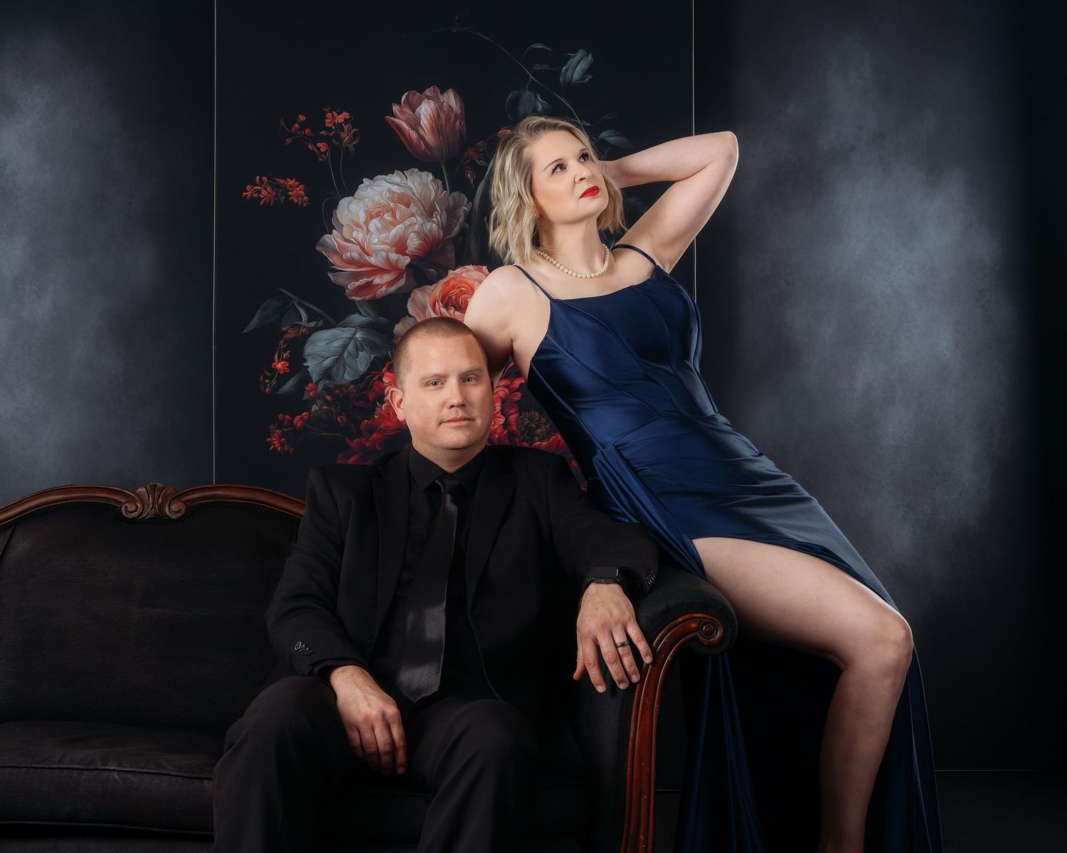 A man in a black suit sits beside a woman in a blue satin dress posing elegantly before a floral backdrop.