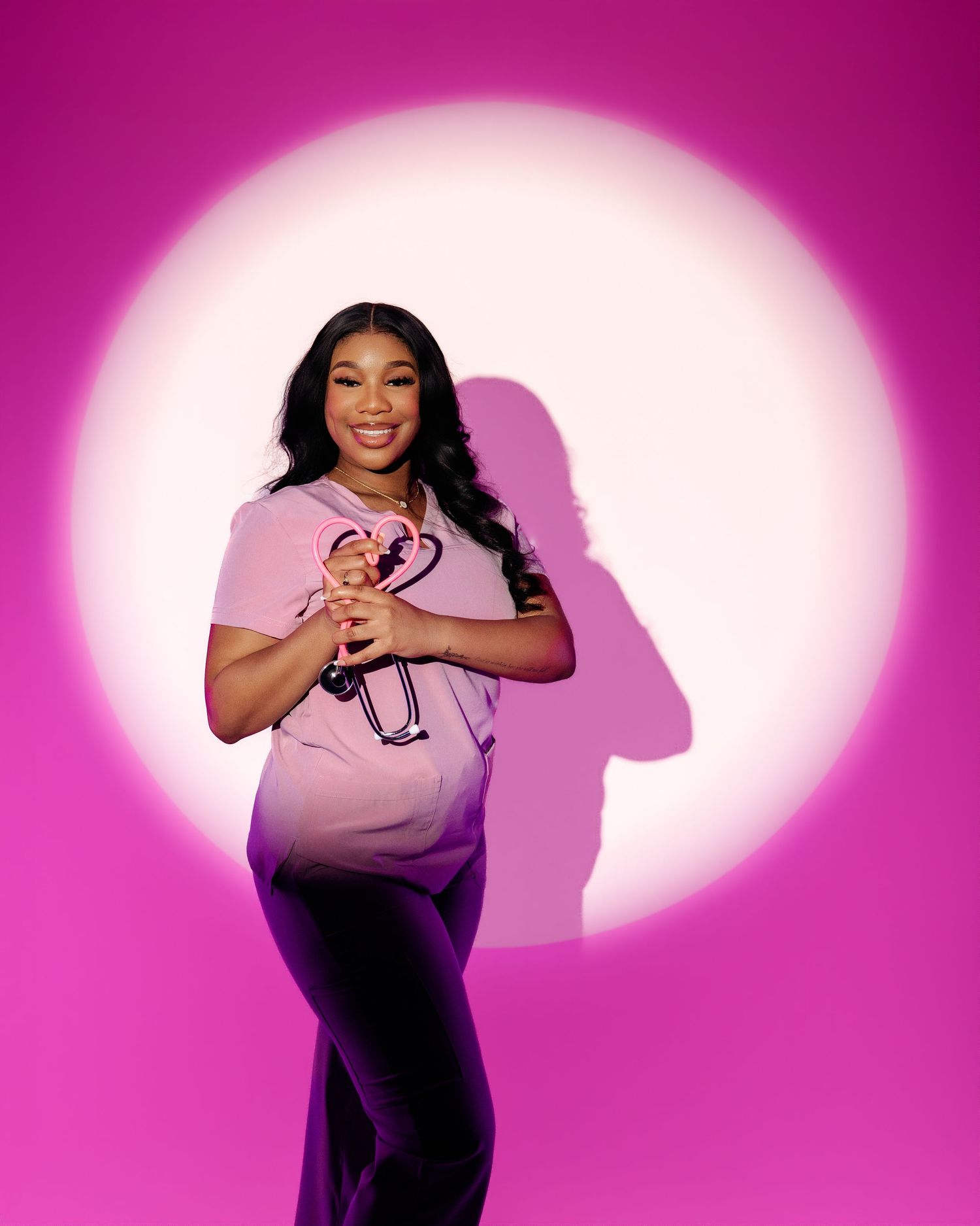 Smiling woman in pink scrubs holding a stethoscope, posed against a vibrant pink background with spotlight effect.