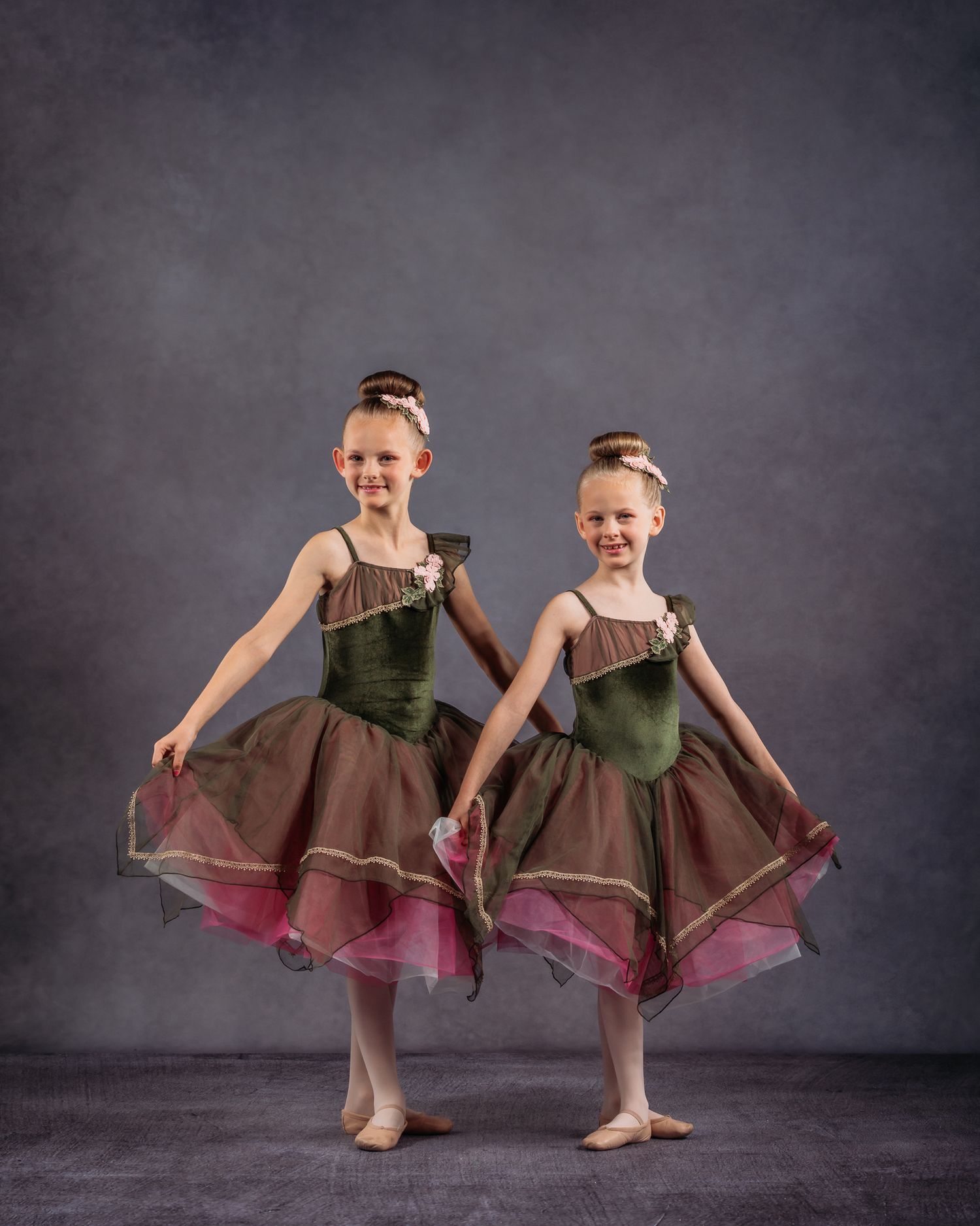 Two young girls in green and pink ballet tutus pose together in a professional dance studio portrait.
