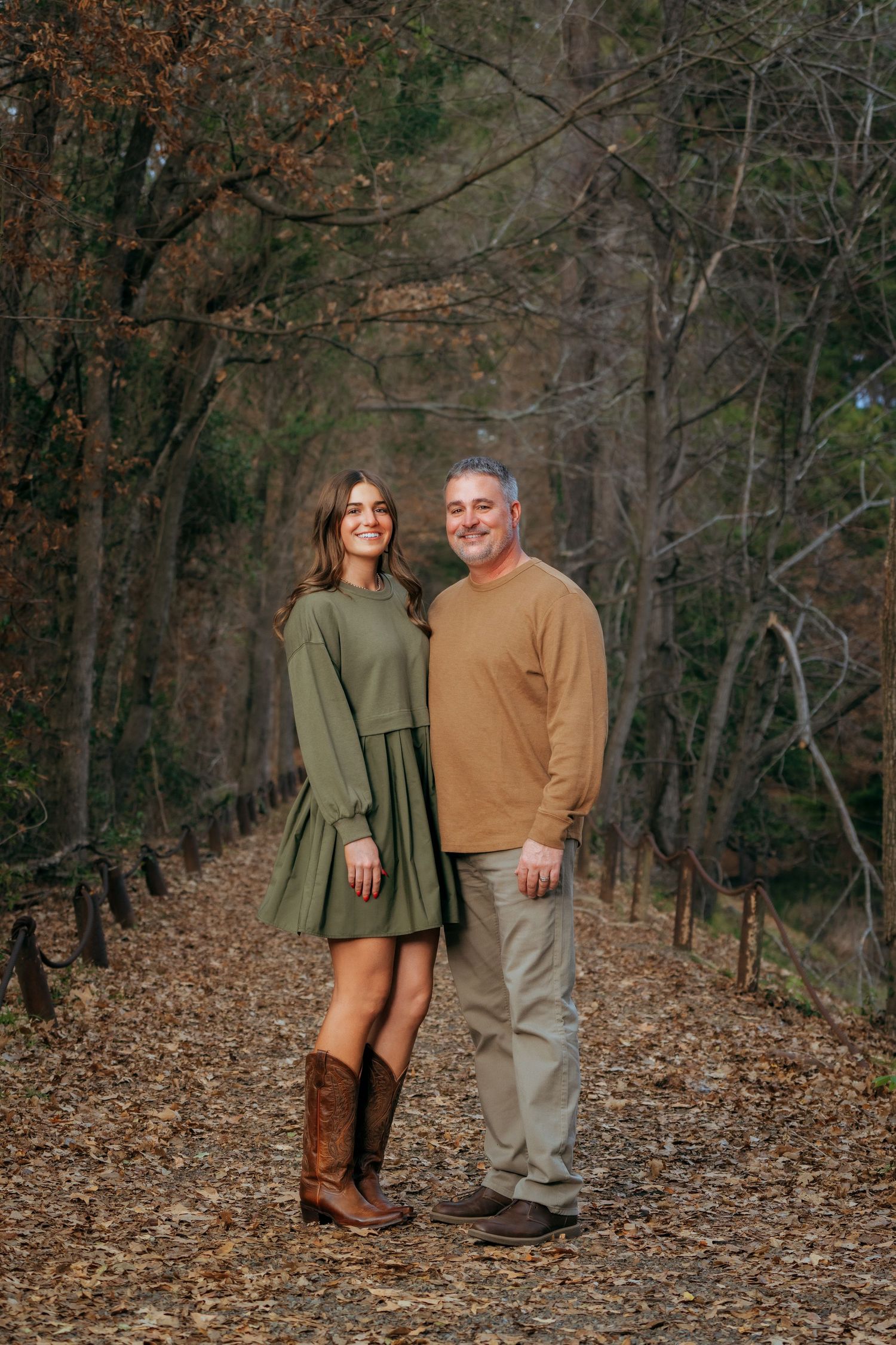 A couple poses on a leaf-covered forest path in autumn, wearing green and tan outfits.