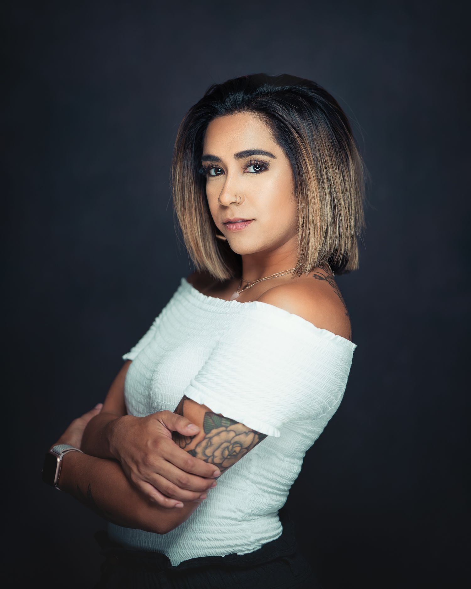 Confident woman with bob haircut and arm tattoo posing with crossed arms against dark backdrop.