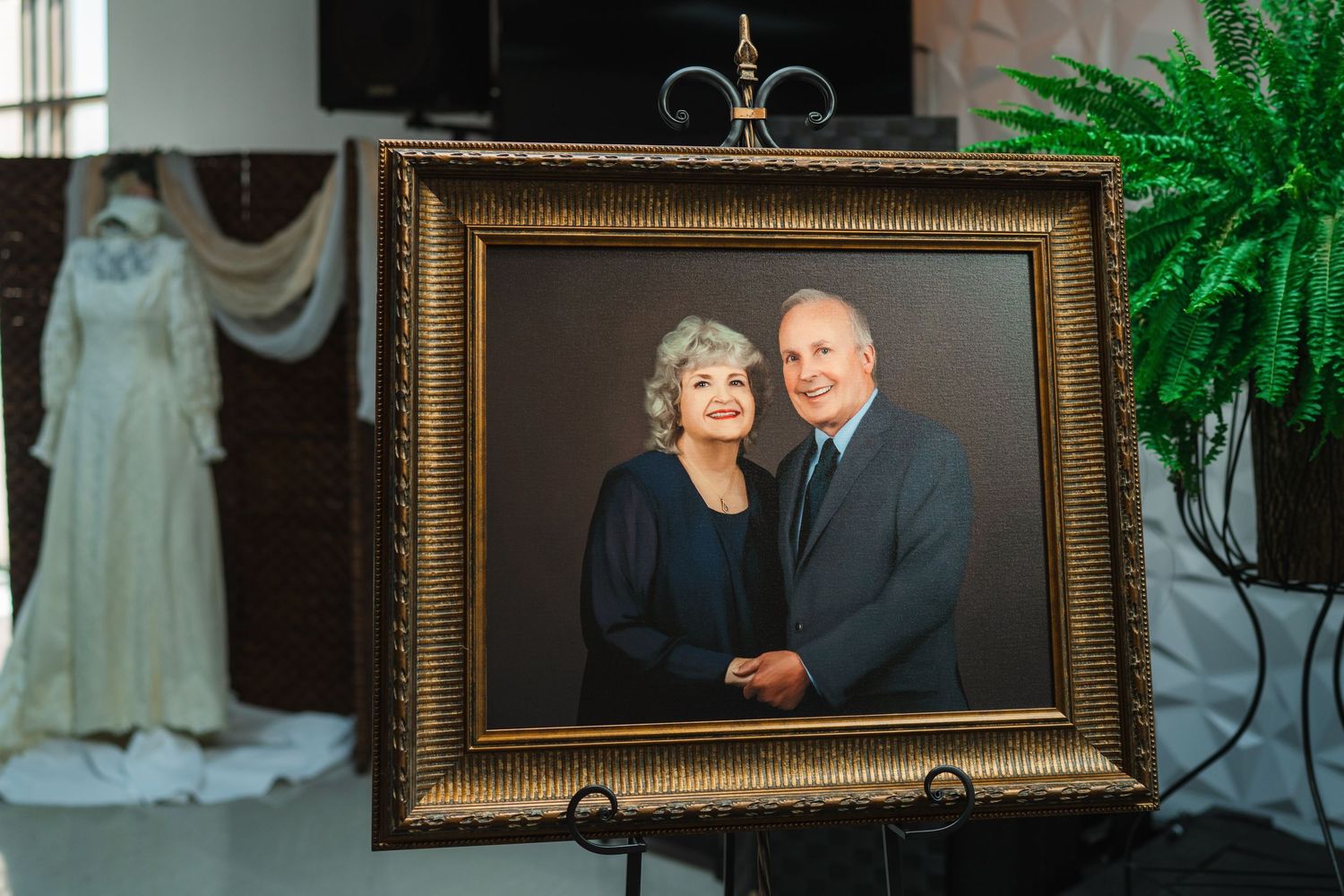 Elegant framed portrait of a smiling older couple displayed on an easel beside a vintage wedding dress.