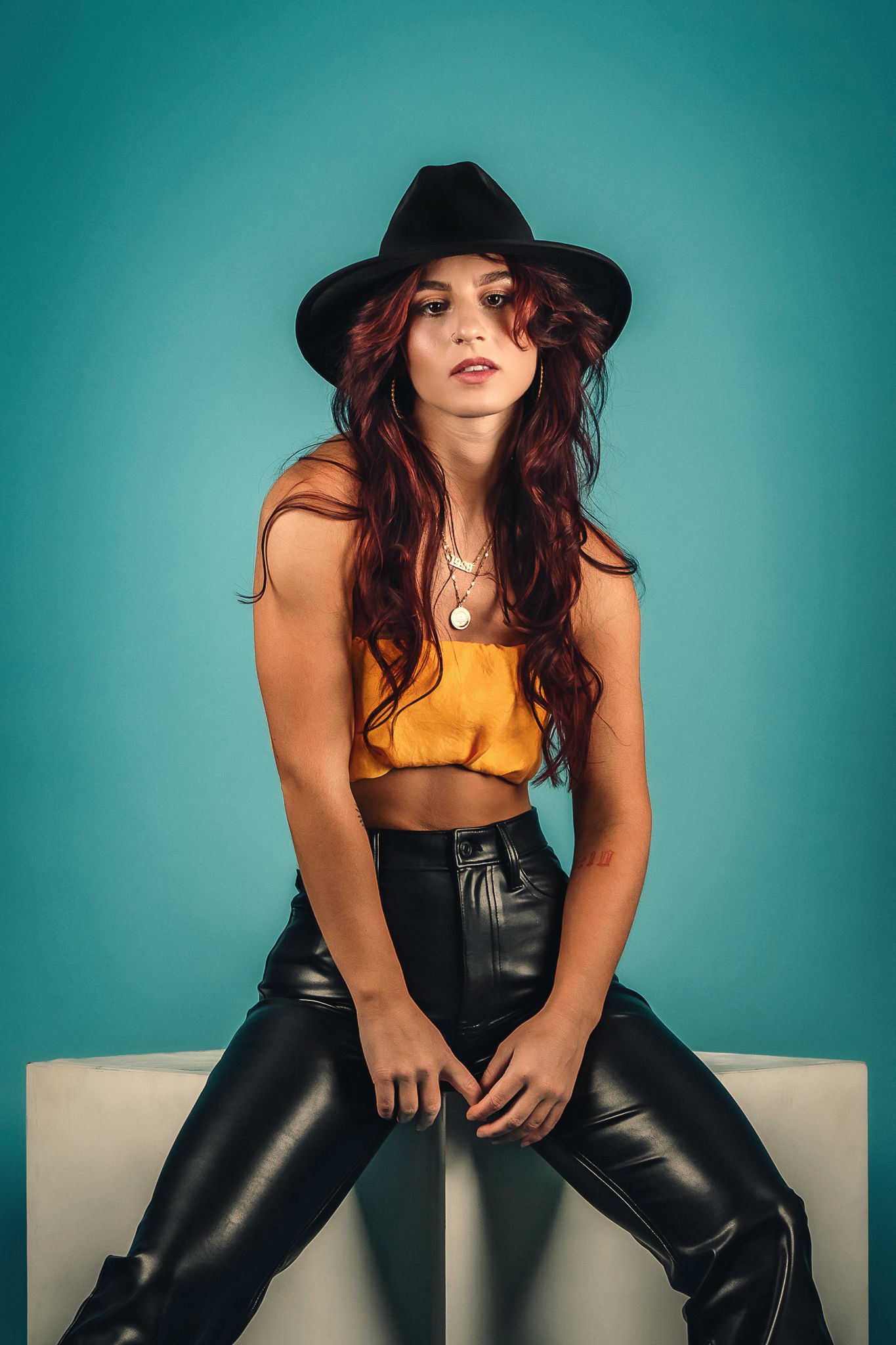 Woman with red hair in black hat and leather pants on teal background.