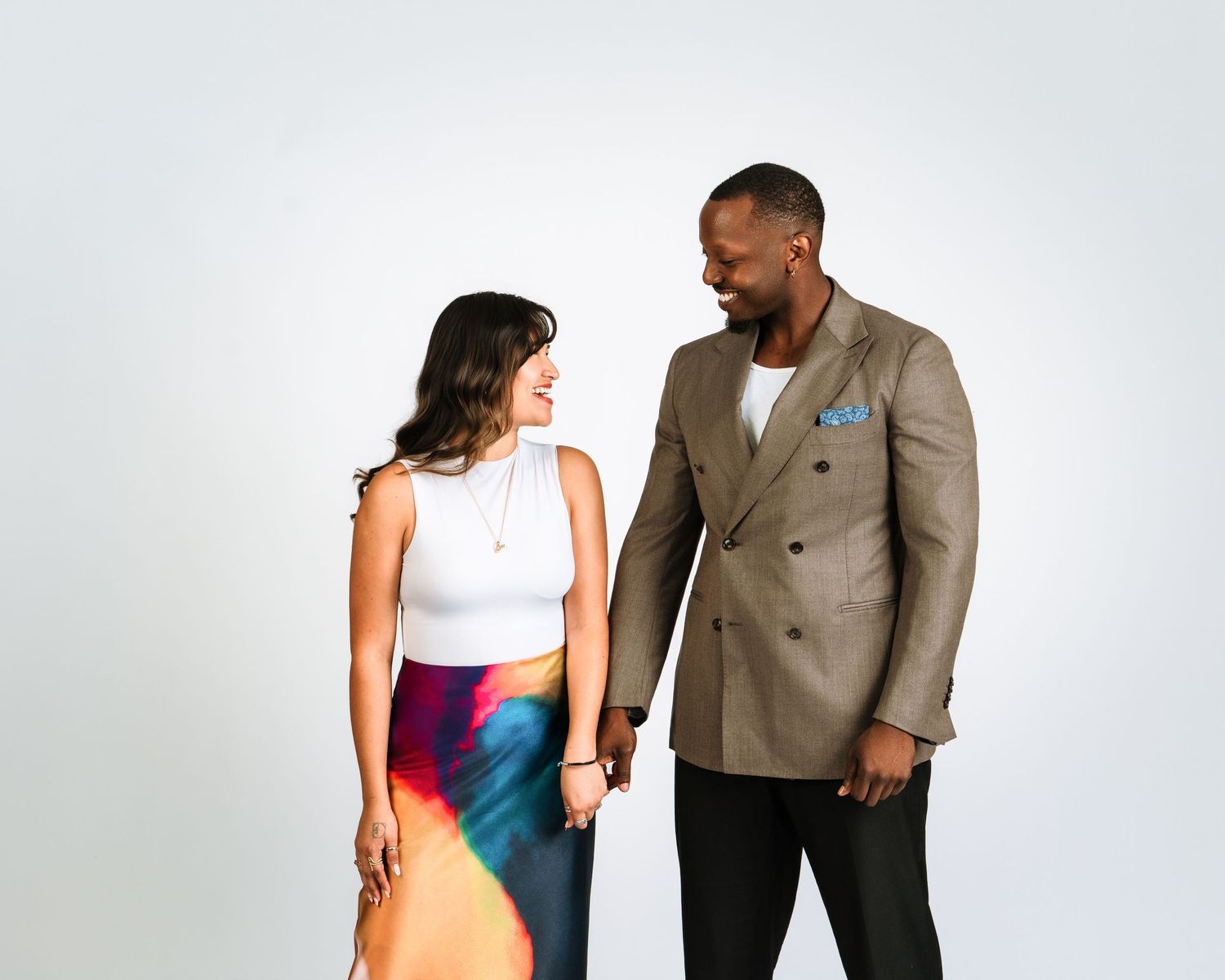 A smiling couple holds hands, the woman in a colorful skirt and white top, the man in a tan blazer.