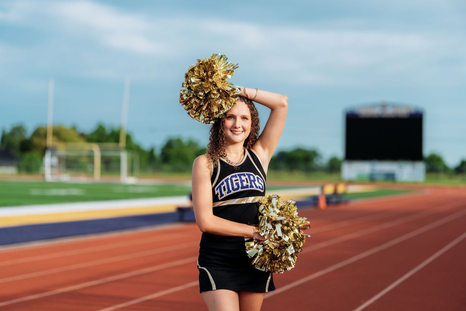 A cheerleader in a black Tigers uniform holds gold pom-poms on a track and field stadium.