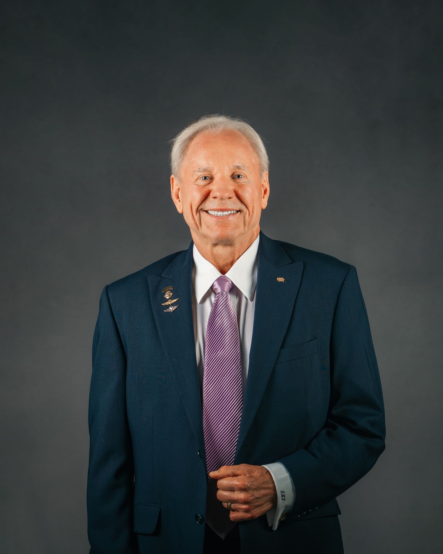 Smiling elderly man in navy suit and purple tie posing for professional headshot portrait.