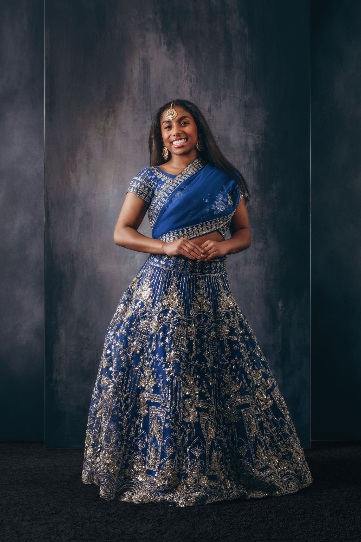 Young woman in ornate blue and gold Indian lehenga with dupatta and maang tikka, smiling against dark backdrop.