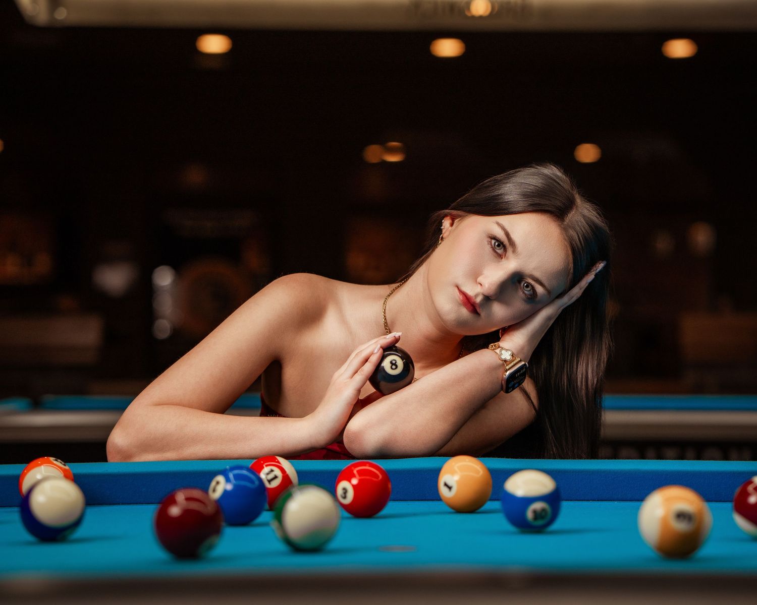 Young woman leaning on a pool table holding a billiard ball, surrounded by colorful billiard balls.