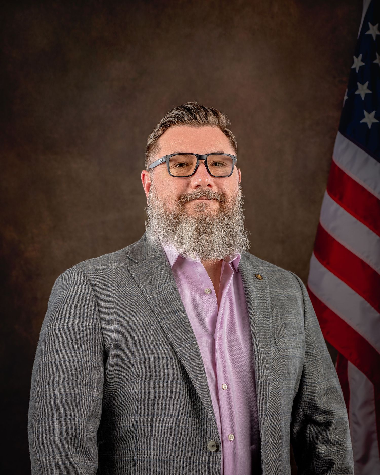 Professional man with beard and glasses in gray blazer poses for formal portrait beside American flag.