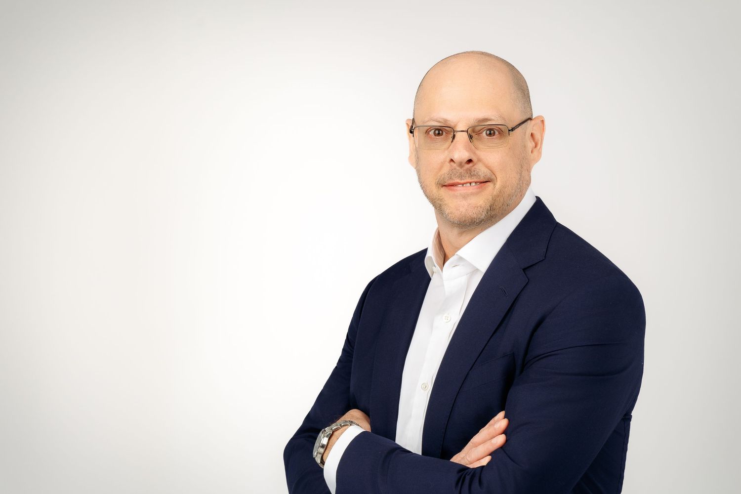 Bald professional man in navy blazer with arms crossed, smiling confidently against a light gray background.