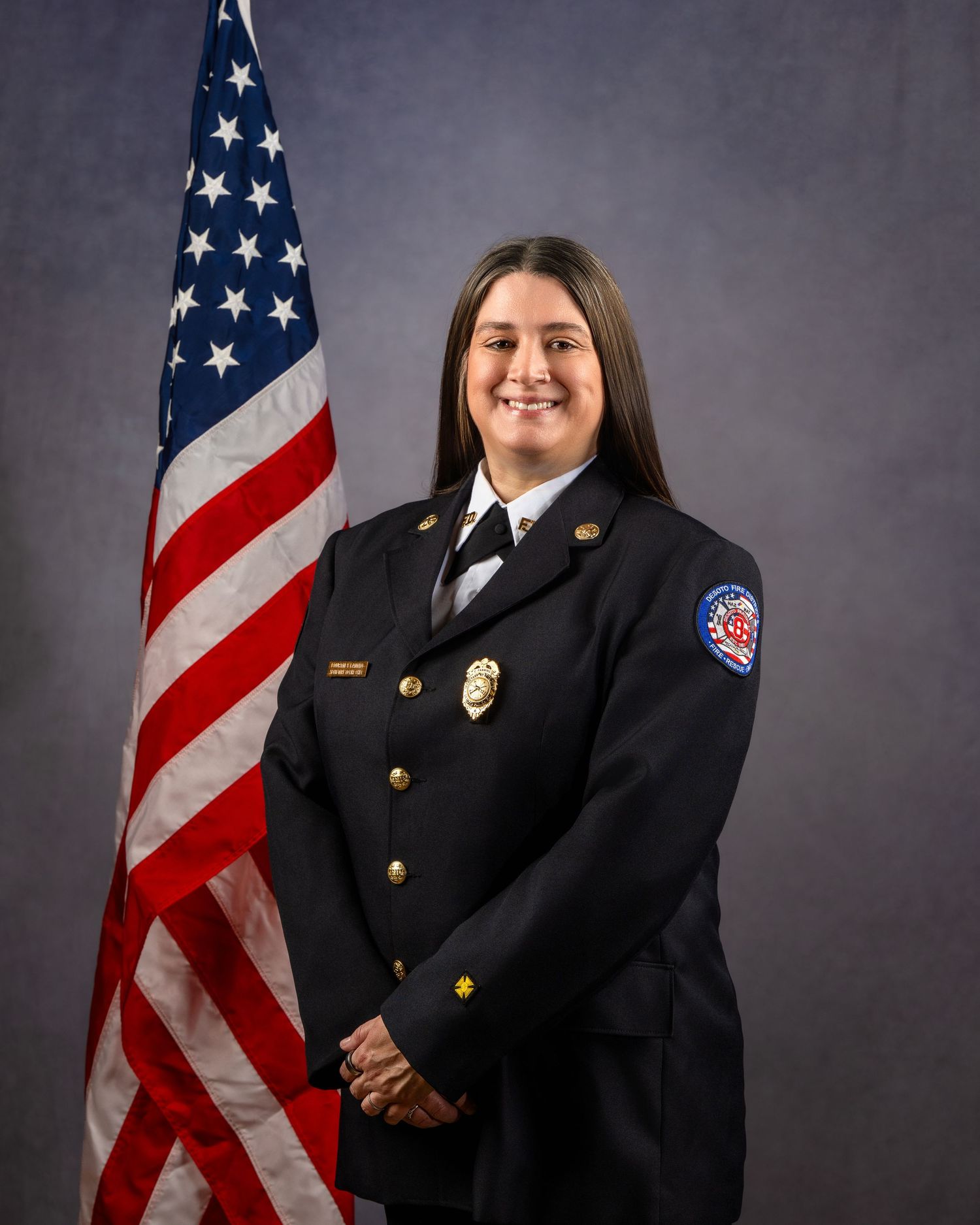 Female fire department officer in dress uniform smiling before American flag in official portrait photo.