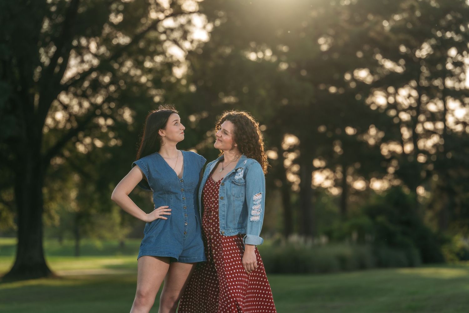 Two young women smiling at each other in a sunlit park with trees in the background.
