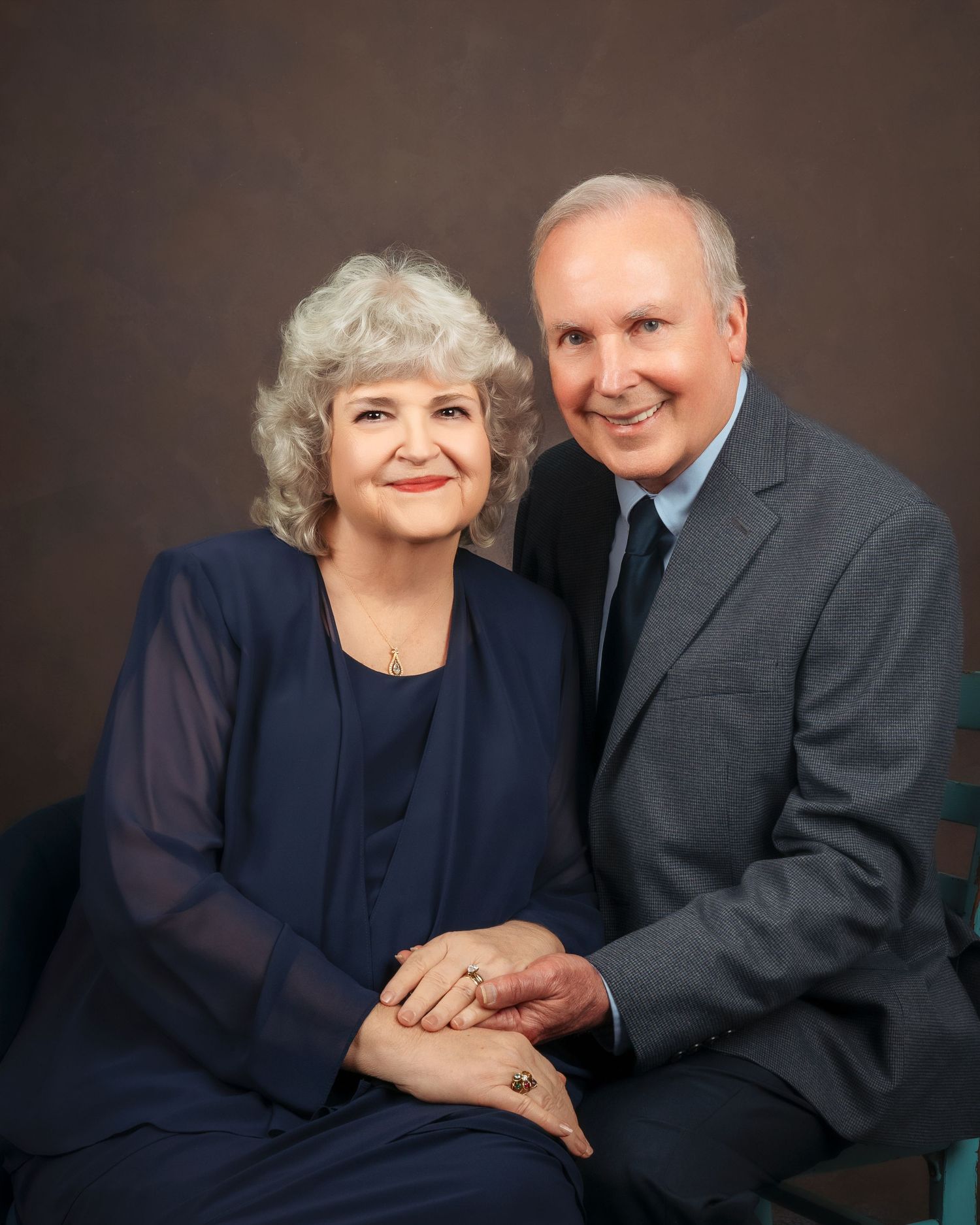 Mature couple smiling warmly in a professional studio portrait, dressed in coordinating navy and gray formal wear.