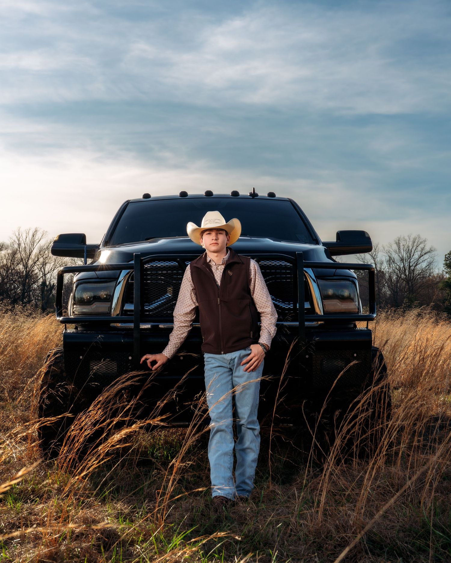 Cowboy in white hat stands before black truck in golden field at dusk.