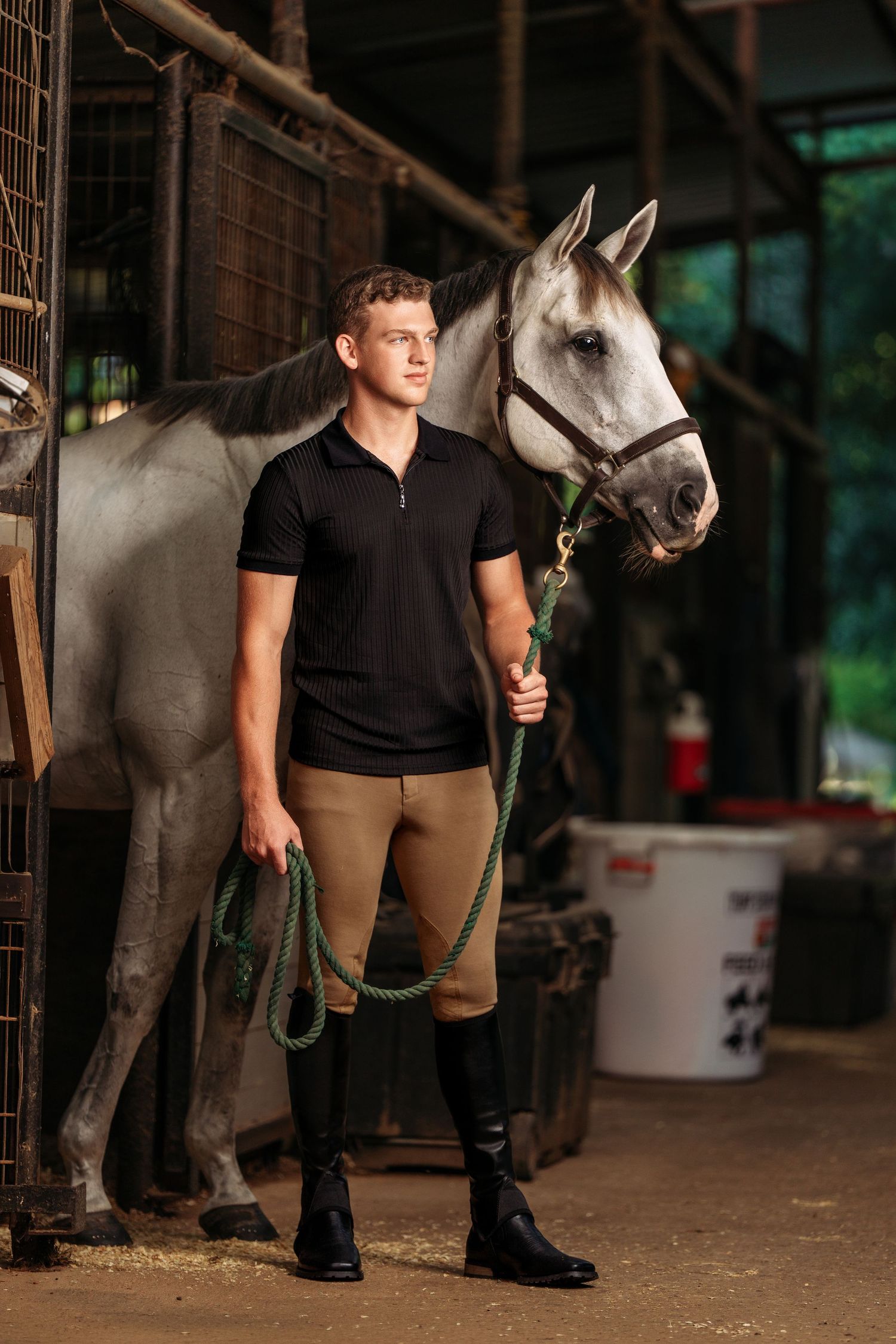 A young man in equestrian attire holds the lead rope of a white horse inside a stable.