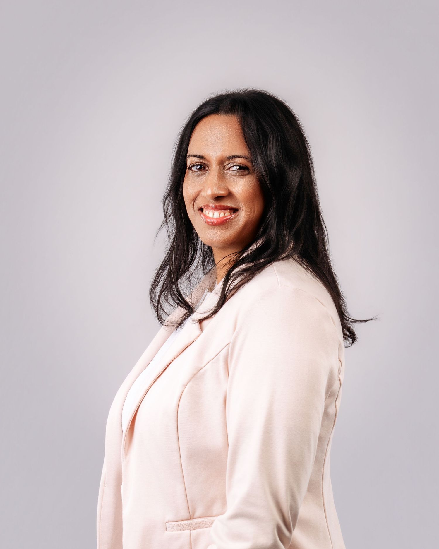 Professional woman in a light pink blazer smiling confidently against a neutral gray background.
