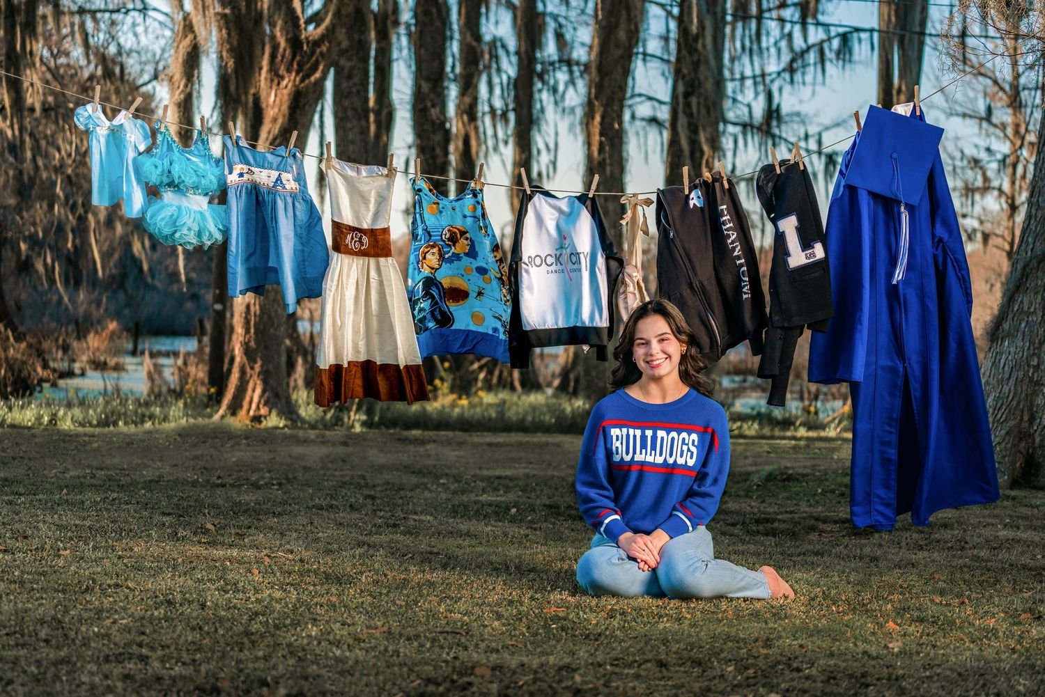 Young girl in Bulldogs sweatshirt sits on grass before clothesline displaying blue and white school uniforms outdoors.