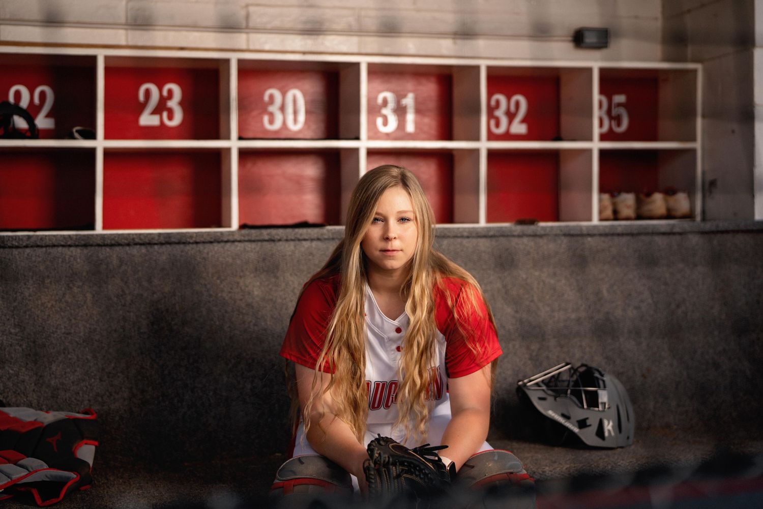 Female softball player sitting in a locker room wearing a red and white uniform with gear.