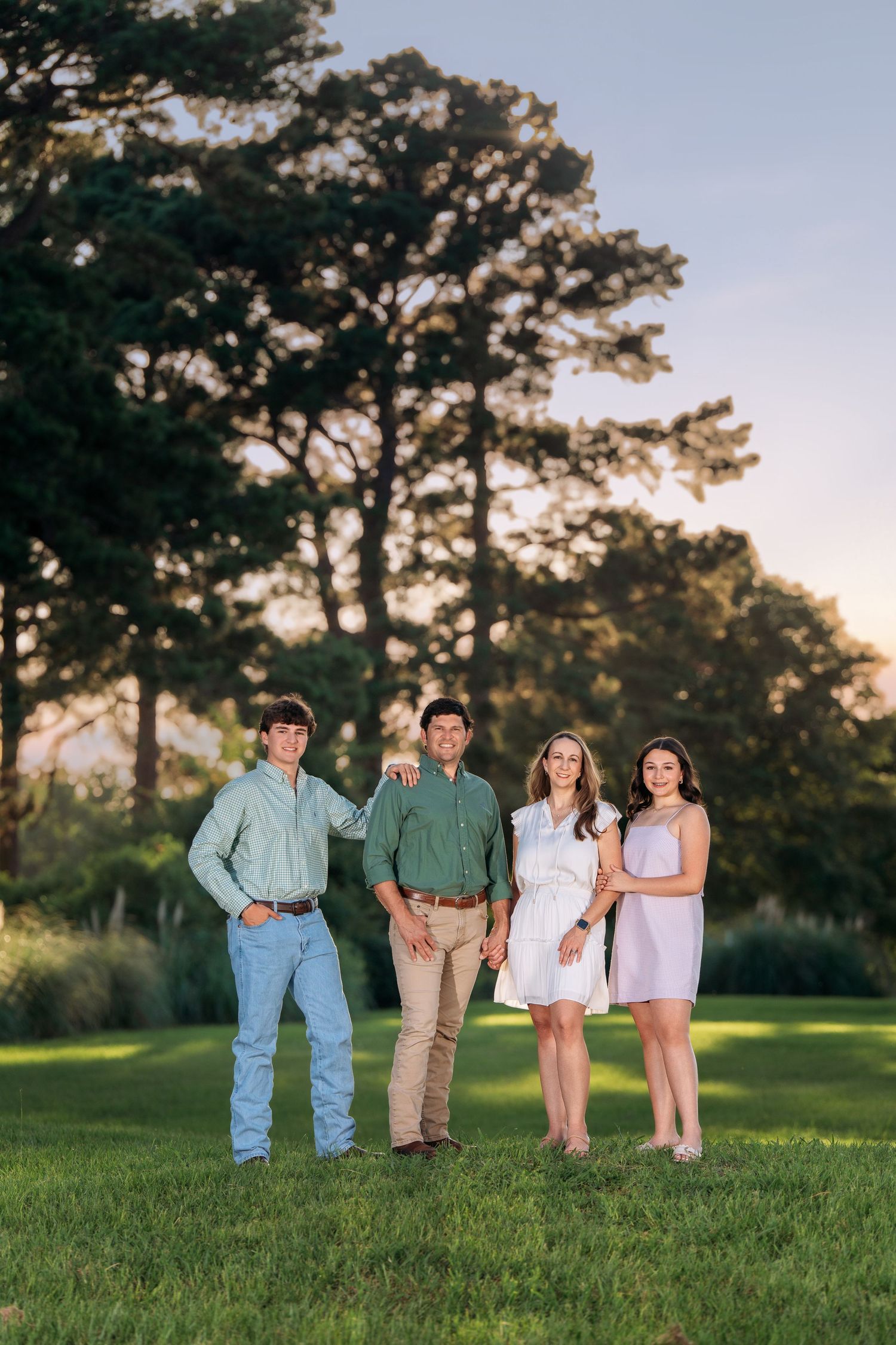 Family of four posing outdoors on green grass with tall pine trees in golden hour sunlight.