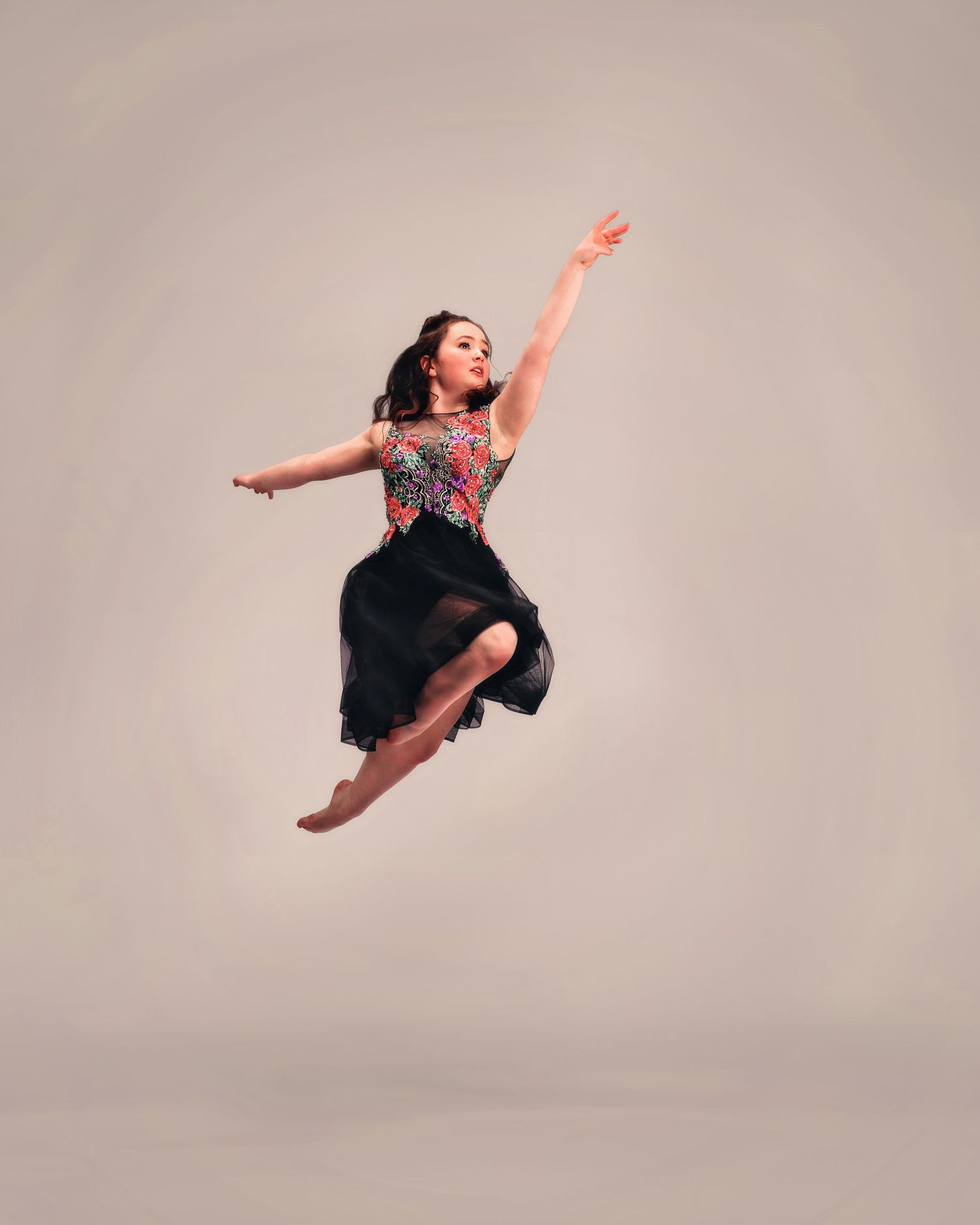 A dancer leaps gracefully in a floral top and black skirt, arms outstretched, captured mid-air against a neutral background.