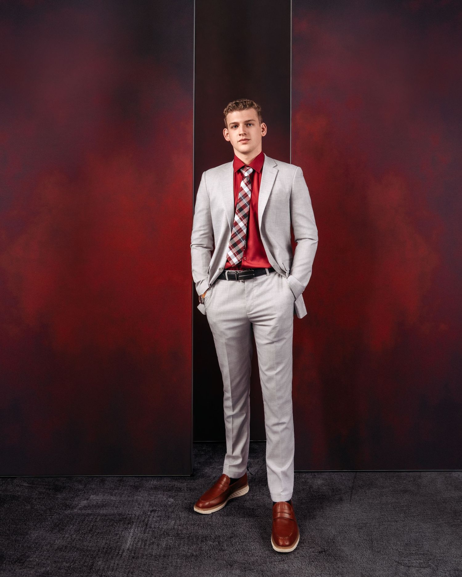 Young man in light gray suit with red shirt and plaid tie, posing against a dark red backdrop.