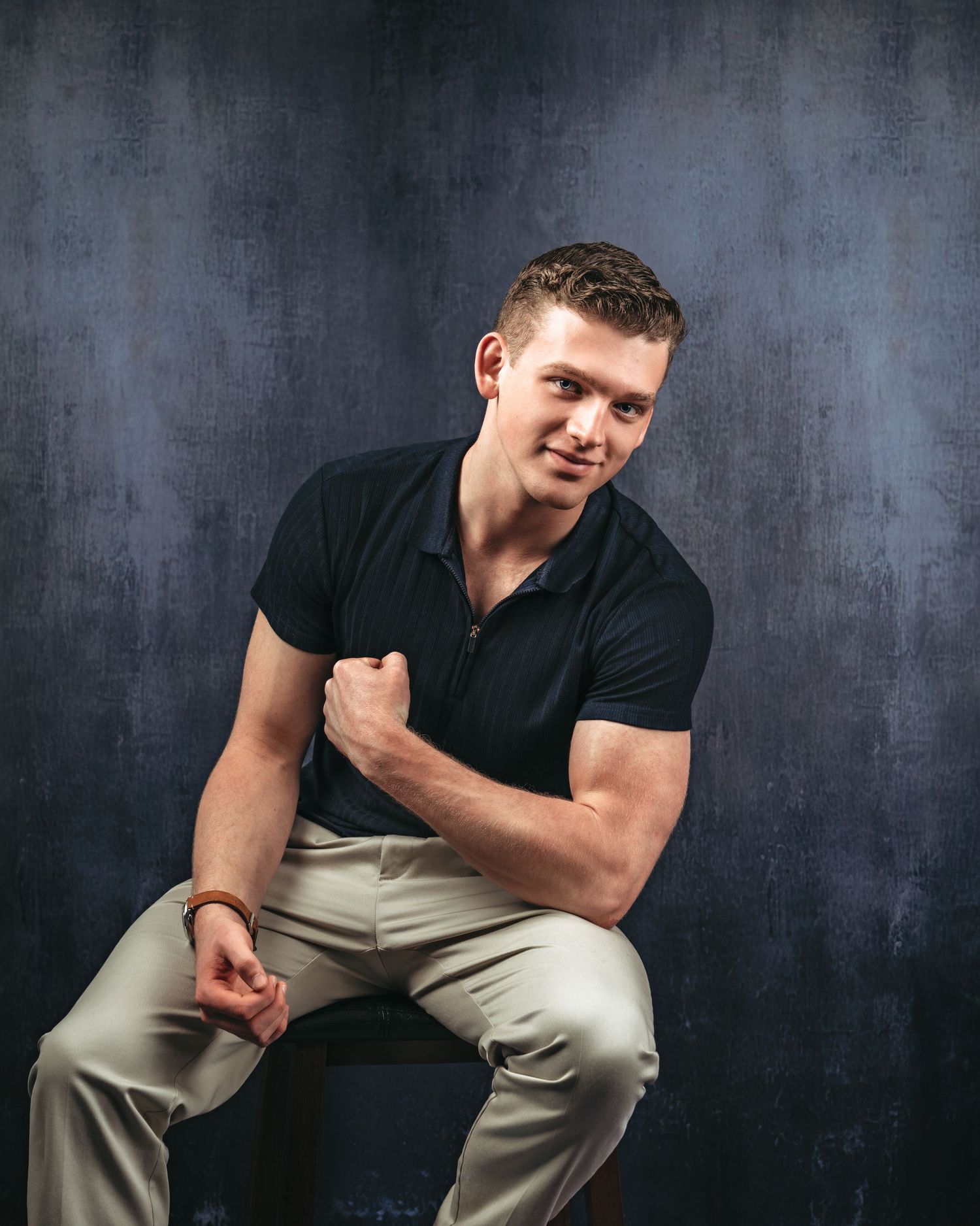 Young muscular man in black polo shirt and khaki pants posing confidently on stool against dark blue textured background.